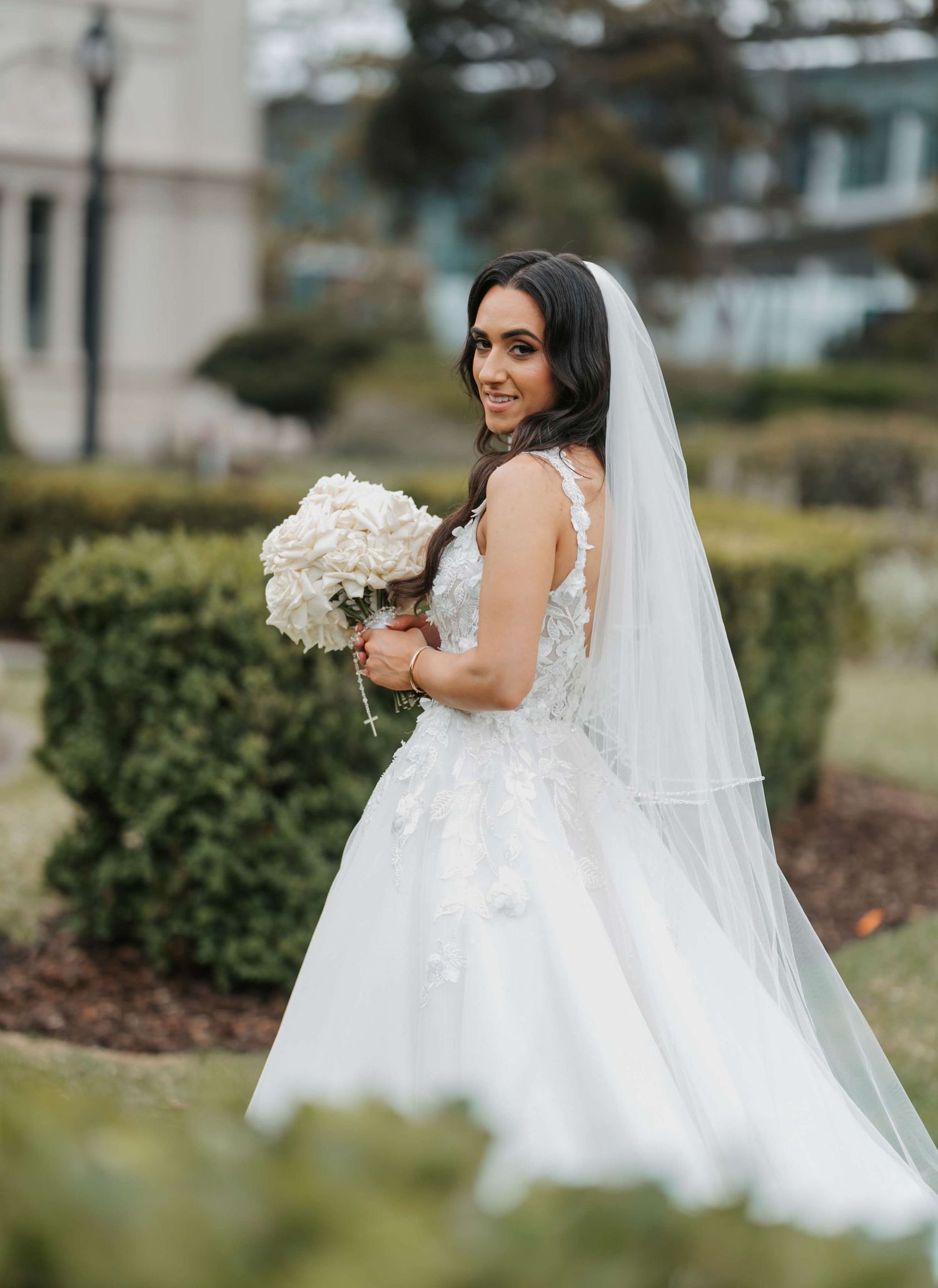 Beautiful bride in white lace wedding gown and long veil holds elegant white rose bouquet in outdoor garden setting.