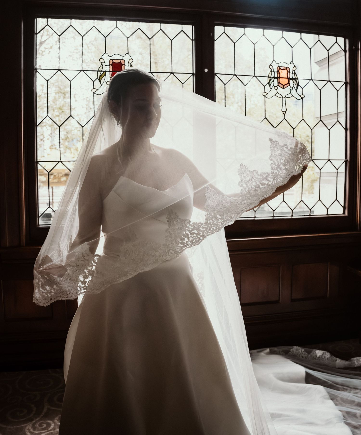 Bride in white wedding gown and lace veil stands by stained glass window in soft backlight.