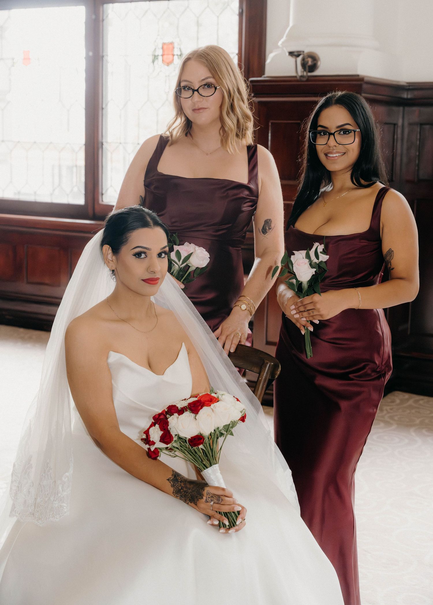 Seated bride in white gown with veil poses with two bridesmaids in dark burgundy satin dresses holding white roses.