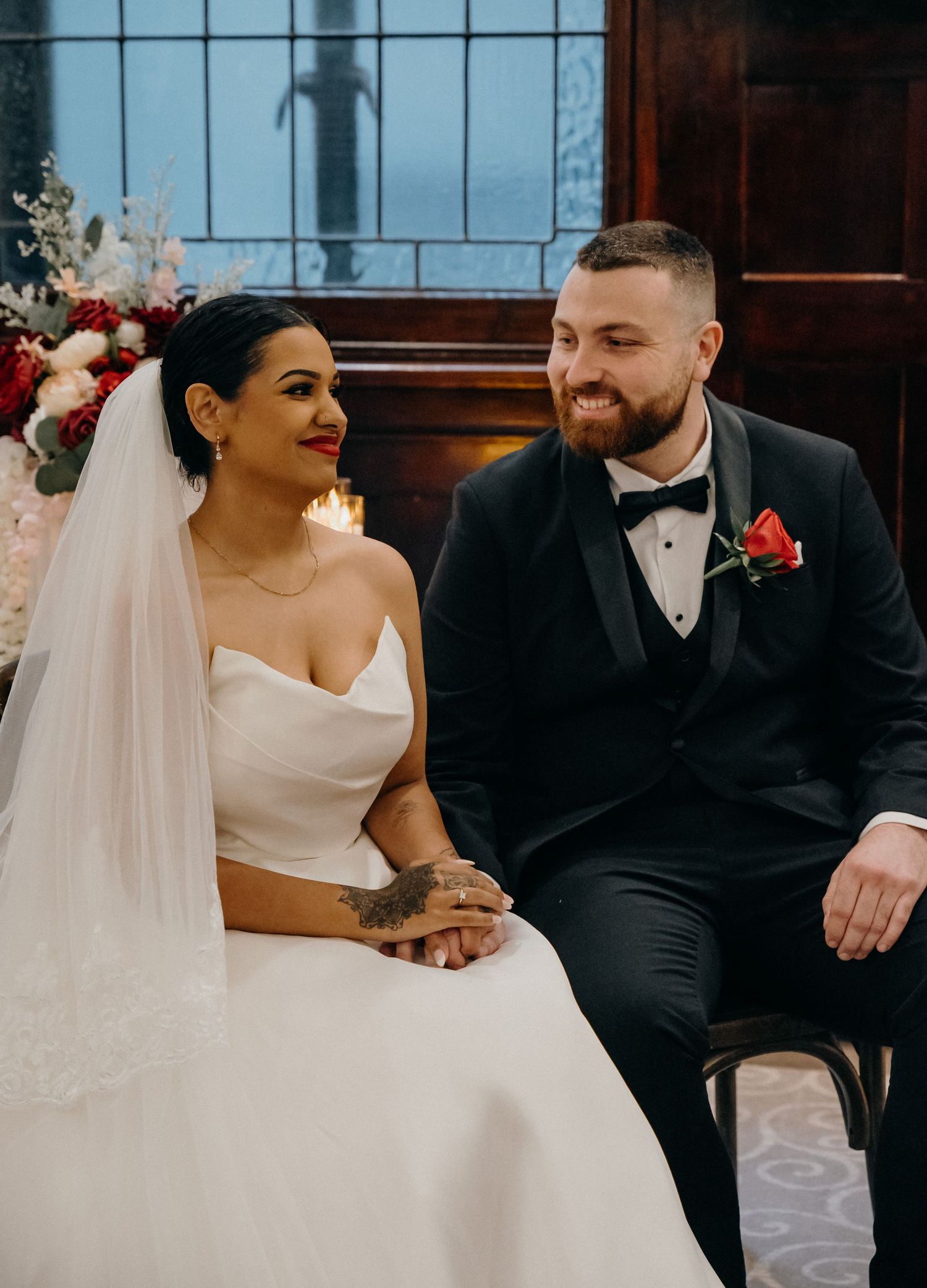Bride in white strapless gown and veil sits beside groom in black tuxedo with red rose boutonniere at wedding ceremony.