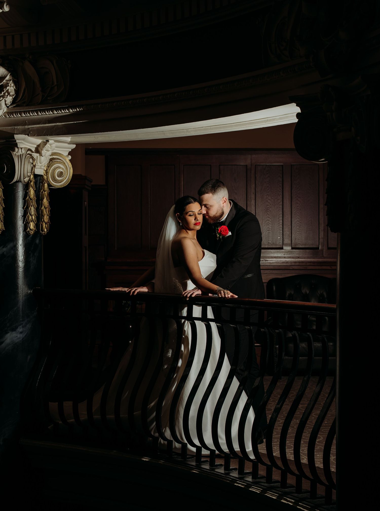 Bride in black and white striped gown with groom in tuxedo embracing on ornate staircase balcony in dramatic lighting.
