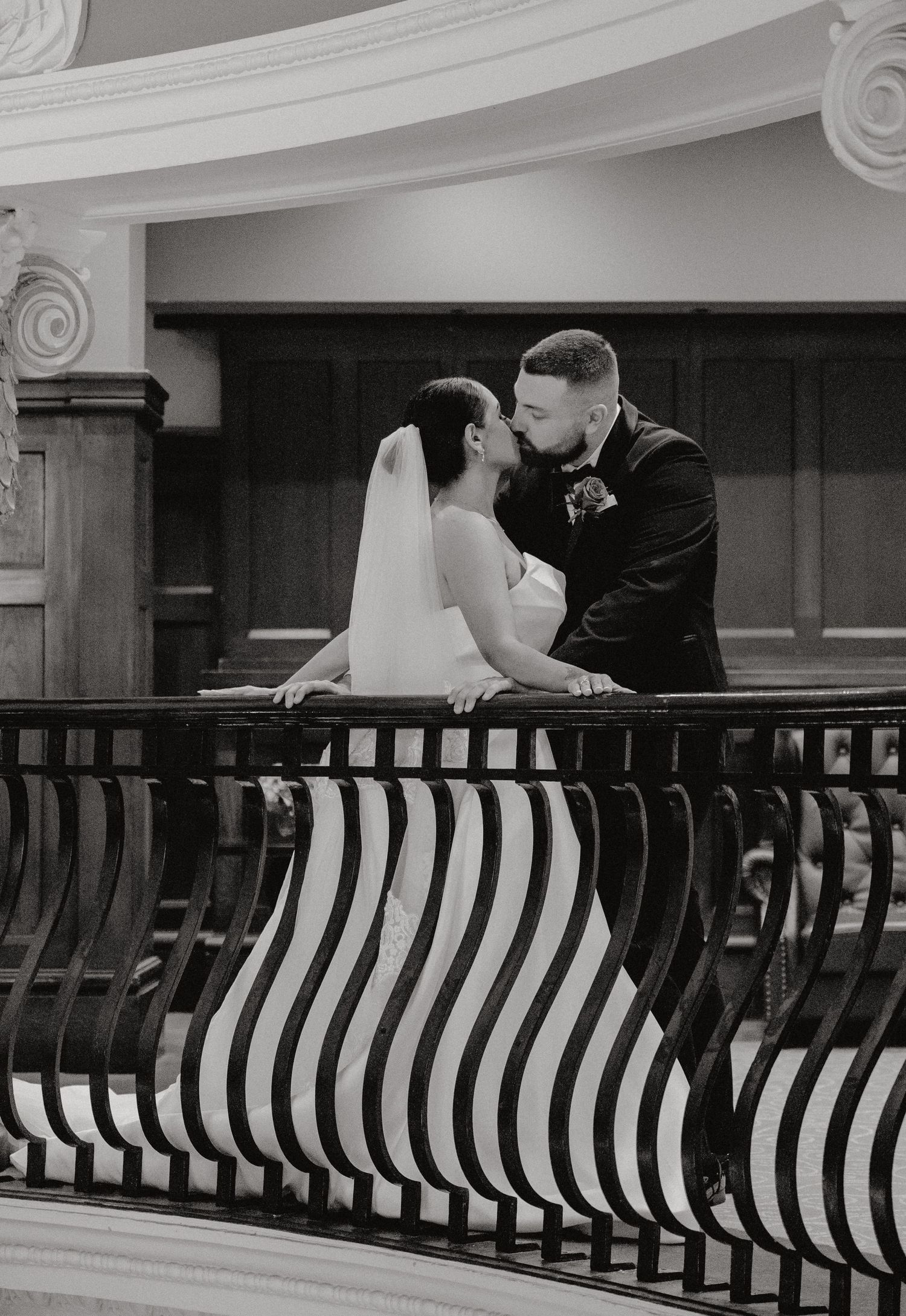Black and white photo of a bride and groom sharing a kiss on an elegant balcony with striped railings.