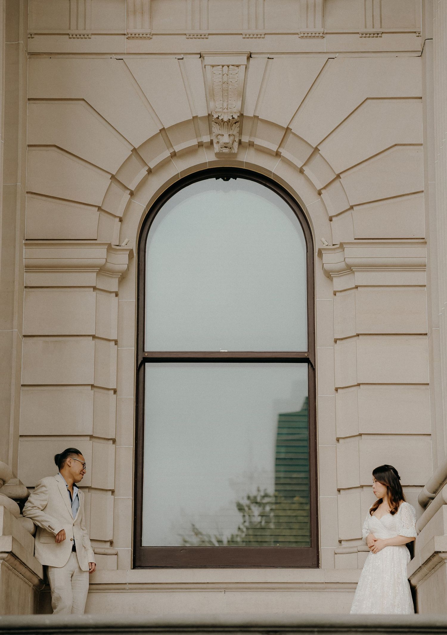 A wedding couple poses in front of an elegant cream-colored building with arched windows and classical architecture.