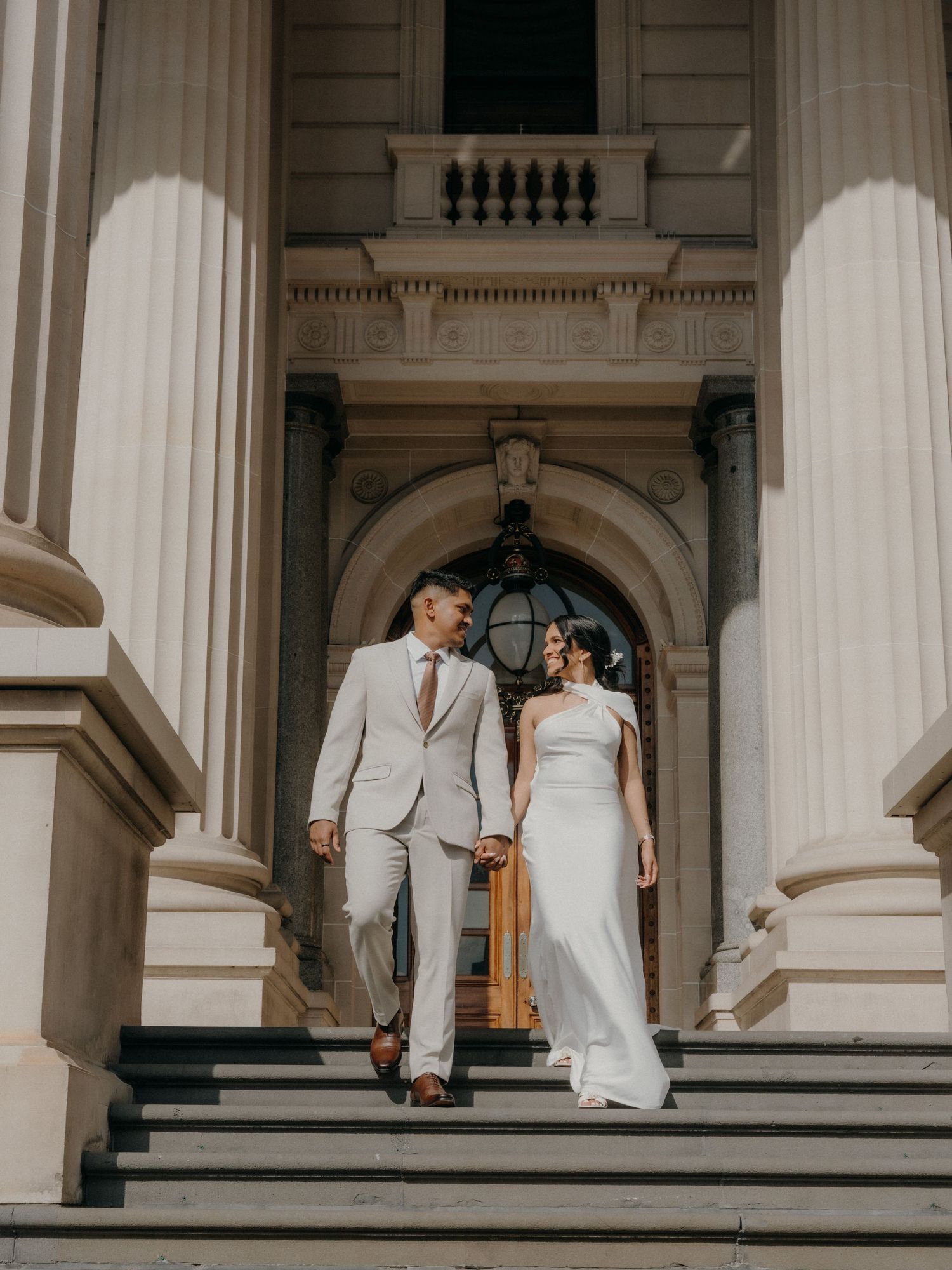 Elegant couple in white wedding attire poses on grand marble steps of historic building with classic columns.