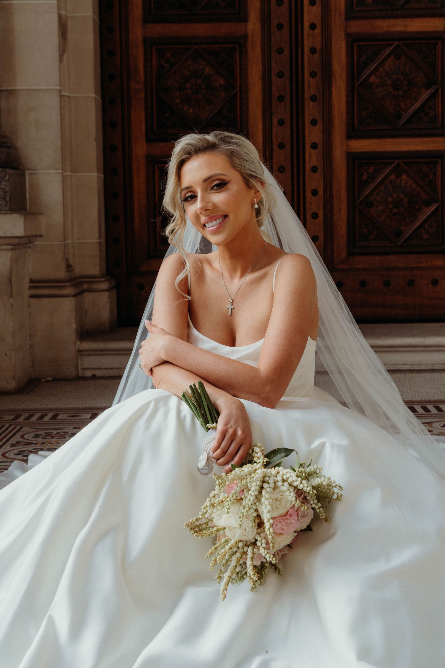 A bride in a white wedding gown poses with her bouquet featuring white and pink flowers against a stone building backdrop.