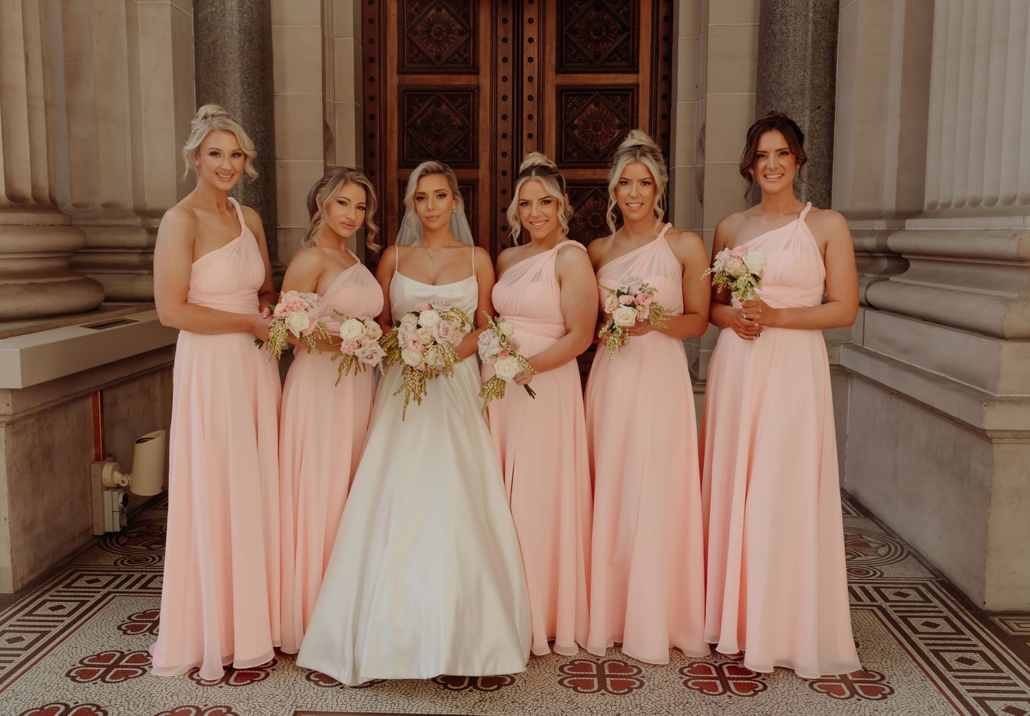 A wedding party wearing pink bridesmaid dresses poses together in front of a grand stone building.
