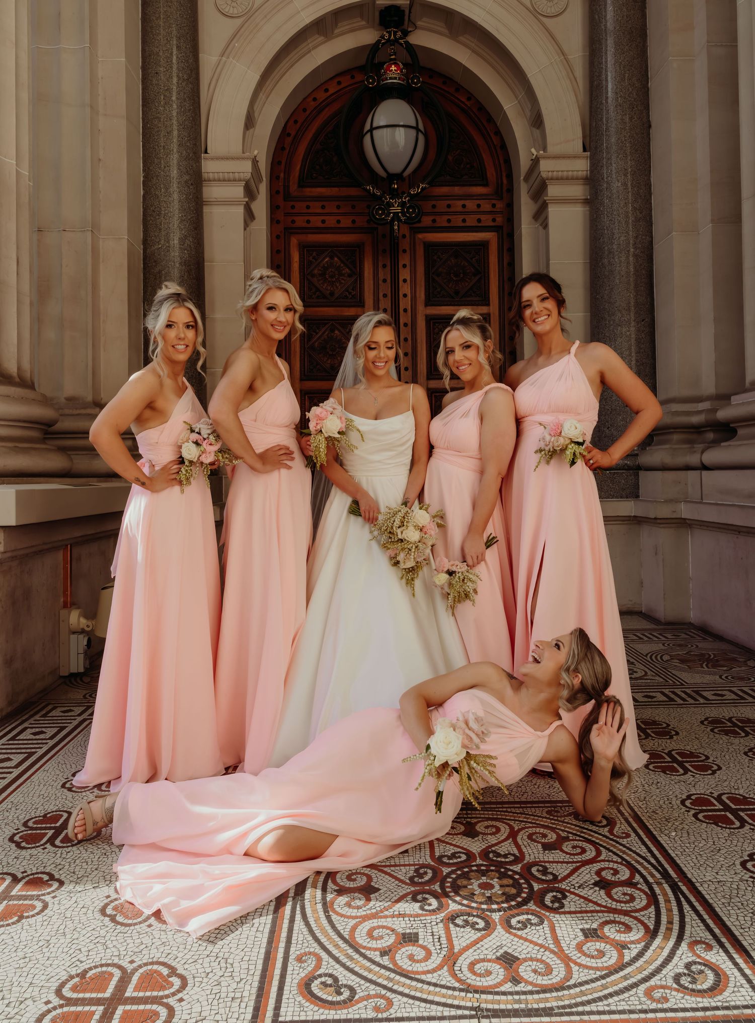 A wedding party in blush pink bridesmaid dresses poses on an ornate tiled floor in an elegant venue.