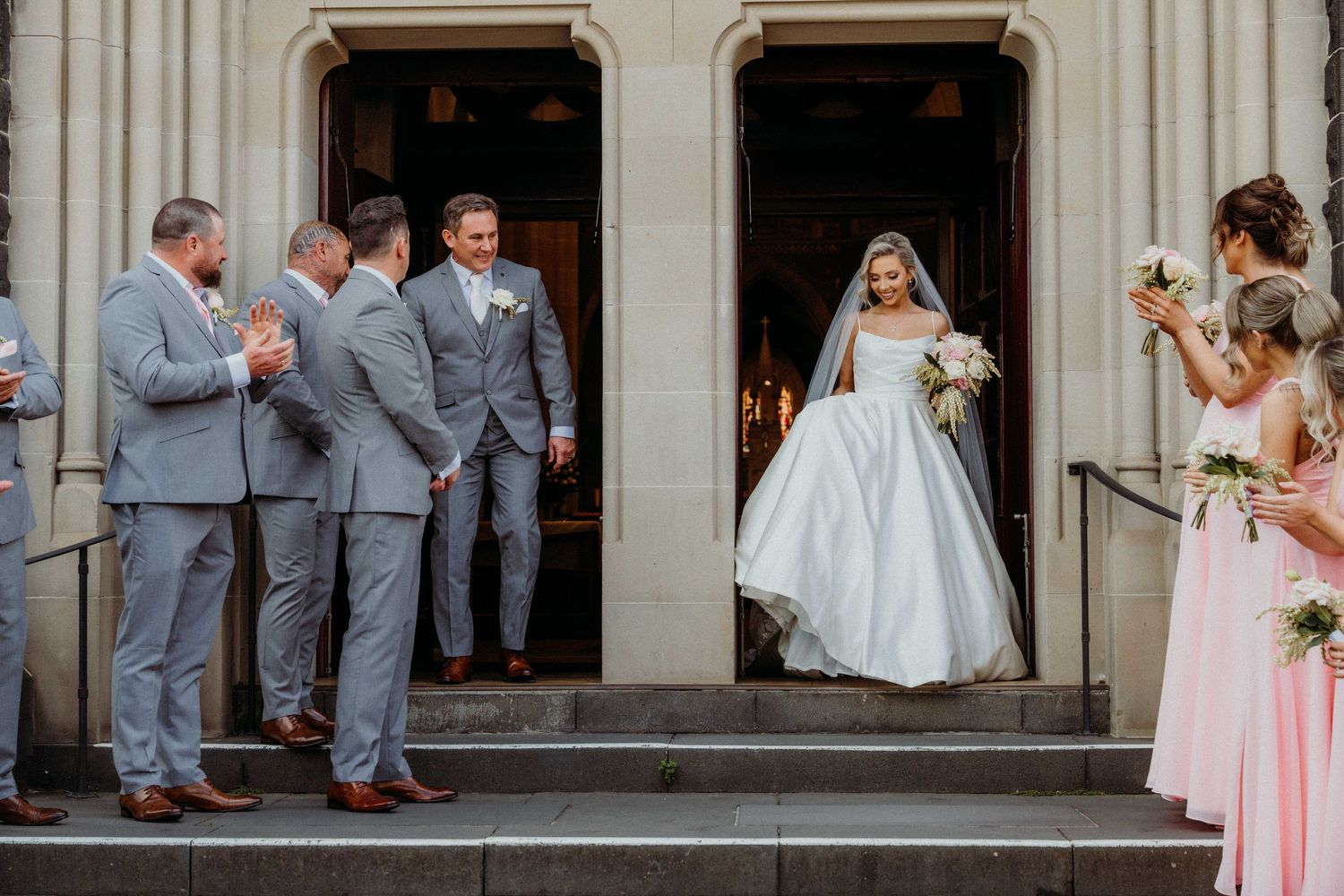 A bride in a white ball gown emerges from church doors while wedding party in gray and pink attire looks on.