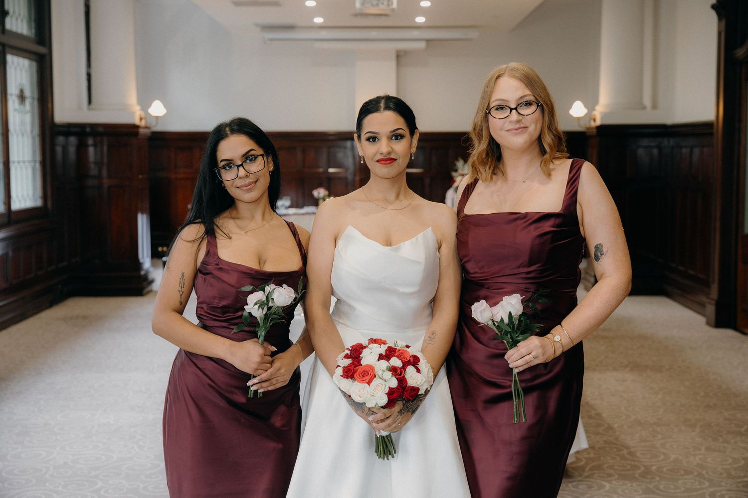 Bride in white gown holds red bouquet flanked by two bridesmaids in burgundy dresses inside elegant venue.