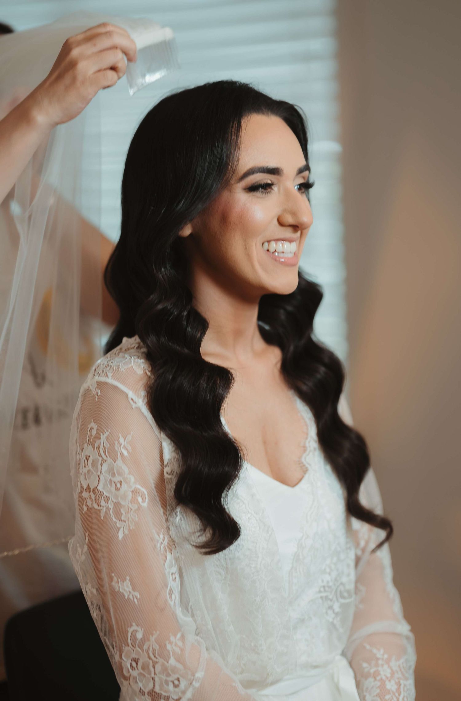 A joyful bride in a white lace wedding dress with long sleeves and dark wavy hair smiles while getting ready.