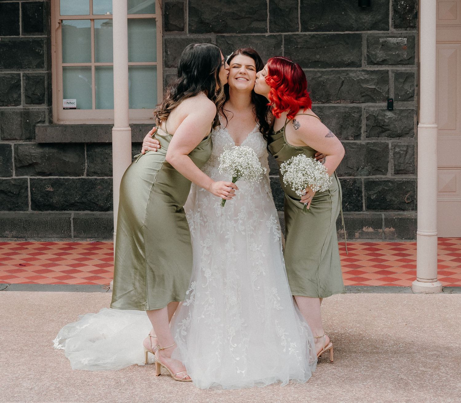 Wedding party members in sage green dresses gather around a bride in a white gown on a stone patio.
