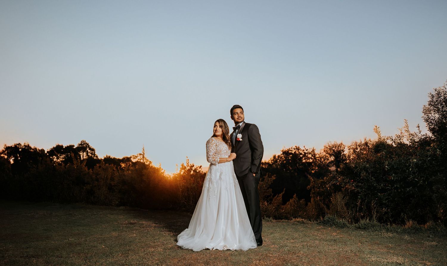A couple in wedding attire poses together on a hilltop at sunset with golden light illuminating the landscape.