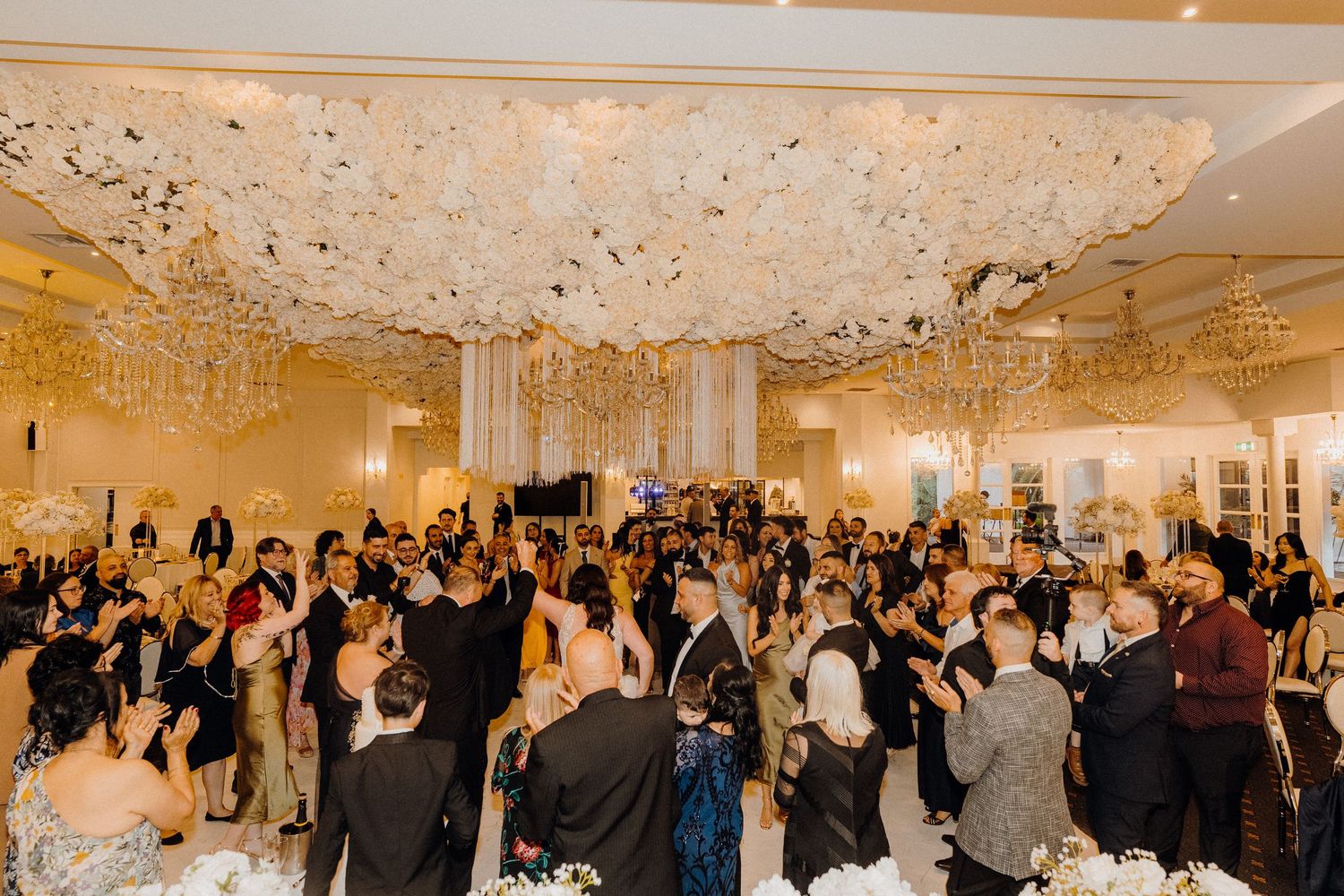 A panoramic view of an elegant wedding reception with guests mingling in a grand ballroom under crystal chandeliers.