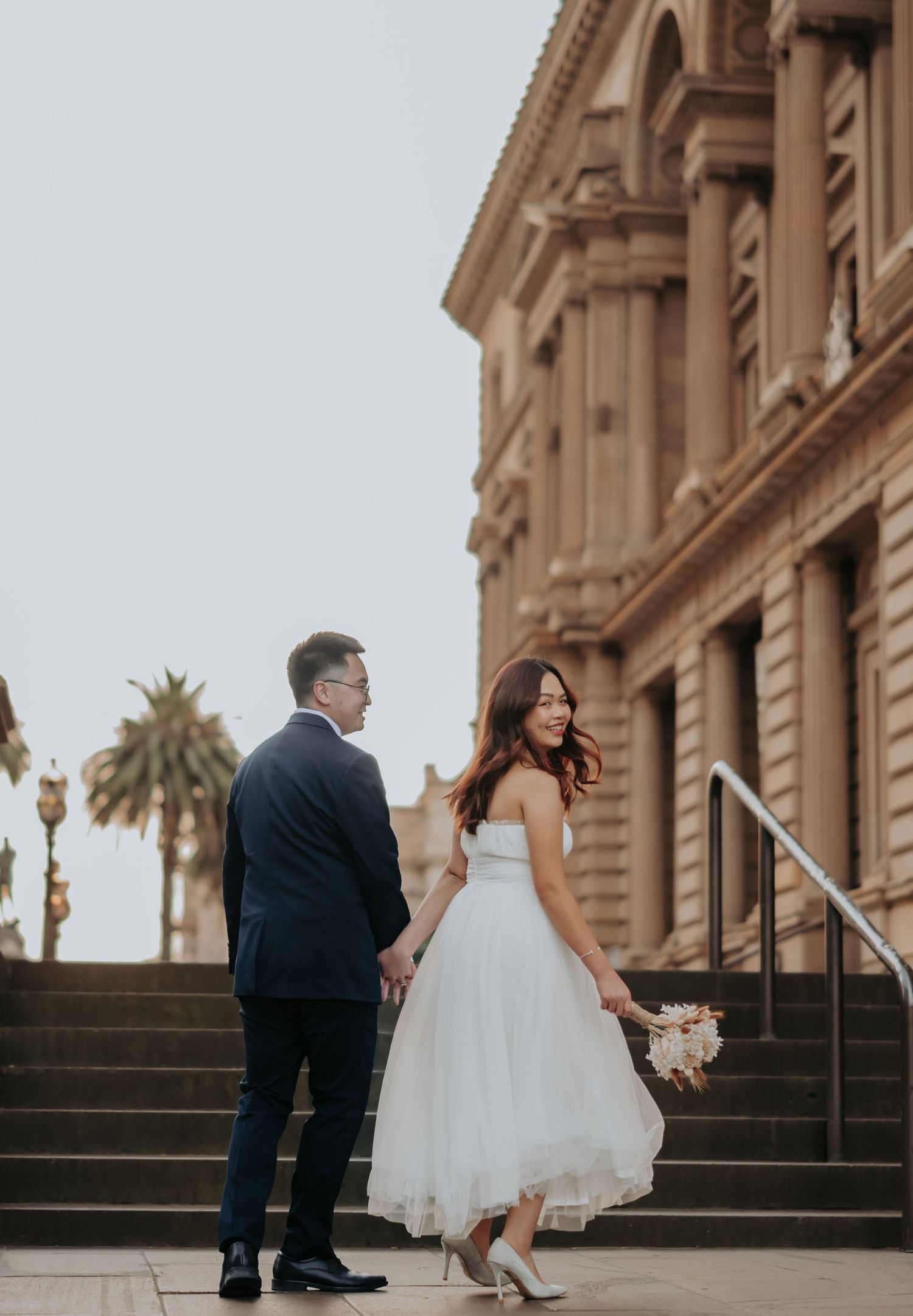 A romantic wedding couple poses on grand stone steps of a historic building at sunset.