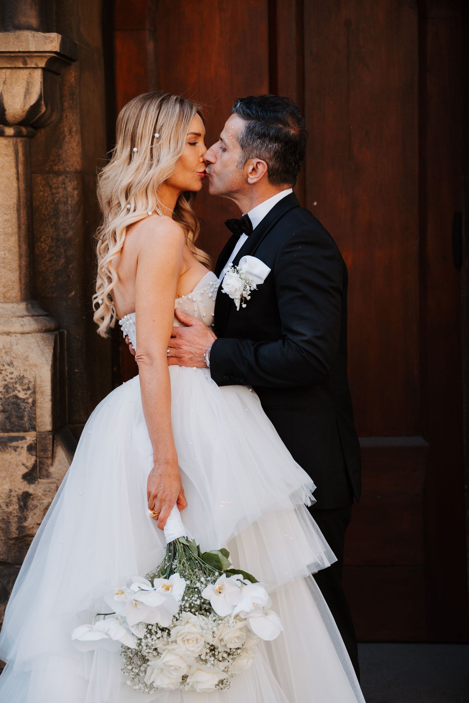 Newlyweds embrace in a tender moment against stone columns while the bride holds a white floral bouquet.