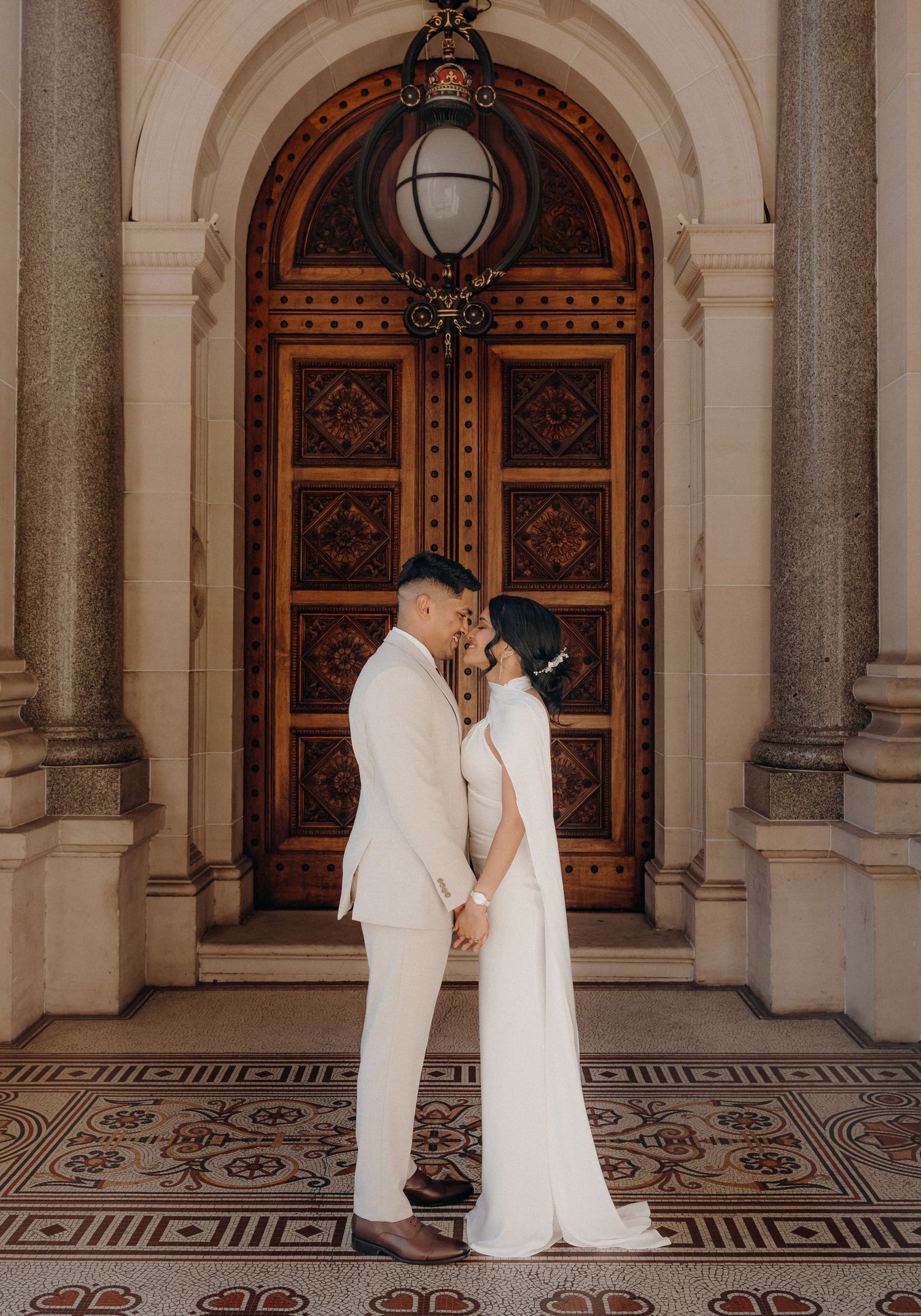 Romantic wedding couple shares intimate moments in an elegant arched hallway with ornate tiled floors and wooden doors.