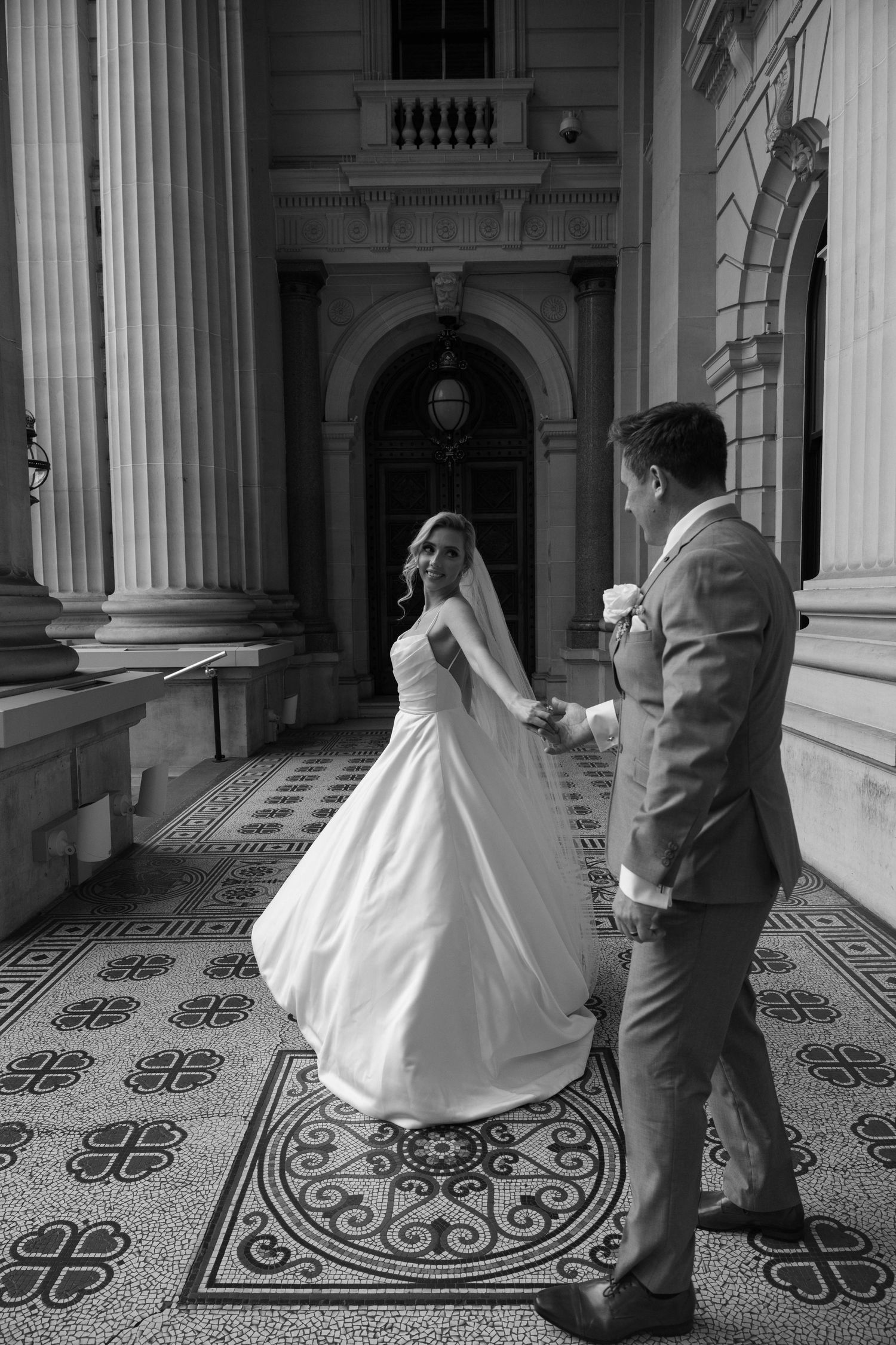 A couple dances in a historic building with ornate tiled floors and classical columns in a black and white photo series.