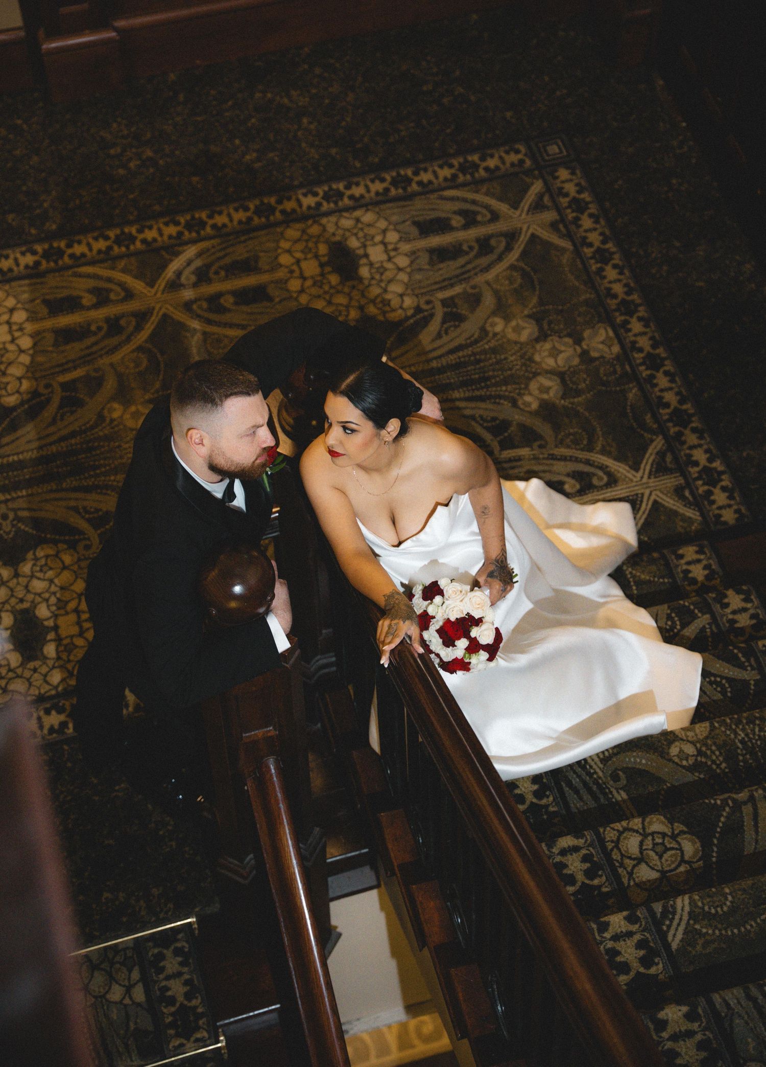 Bride in white gown and groom in black suit share a romantic moment on ornate staircase, bride holding red bouquet.