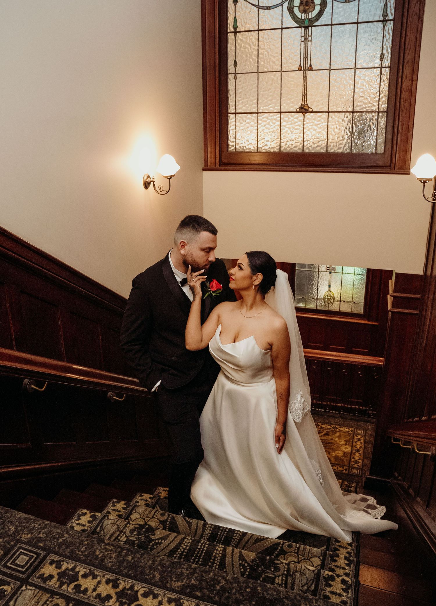 Bride and groom share romantic moment on ornate staircase, bride in strapless gown with veil, groom in black suit.