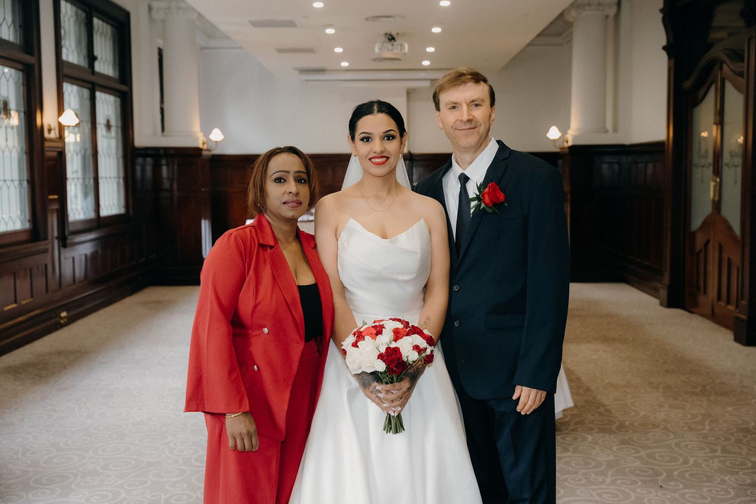 Bride in white gown holding red bouquet poses with groom in suit and woman in red jacket at elegant wedding venue.