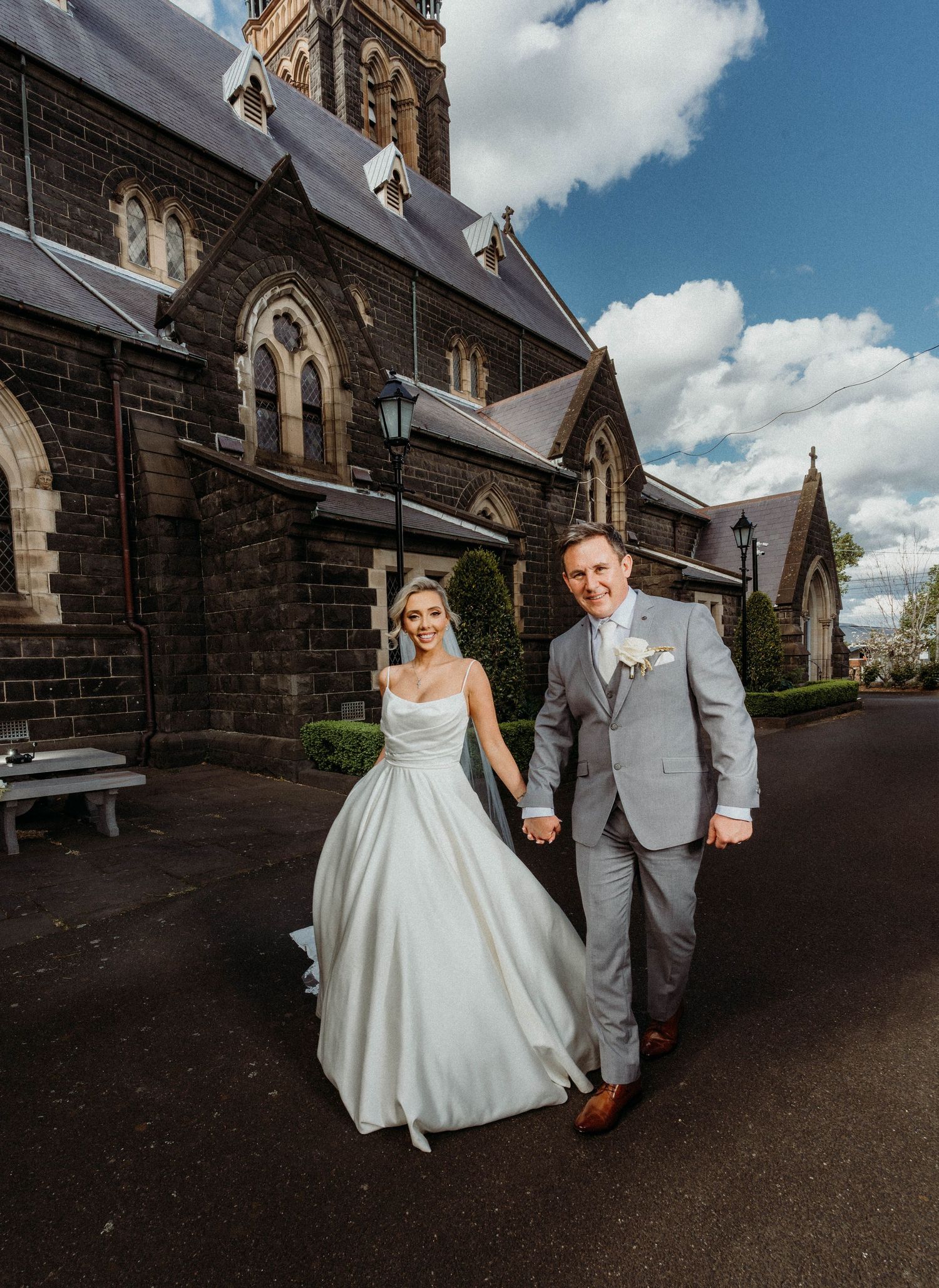 A couple in wedding attire walks hand in hand outside a grand Gothic-style stone church on a sunny day.