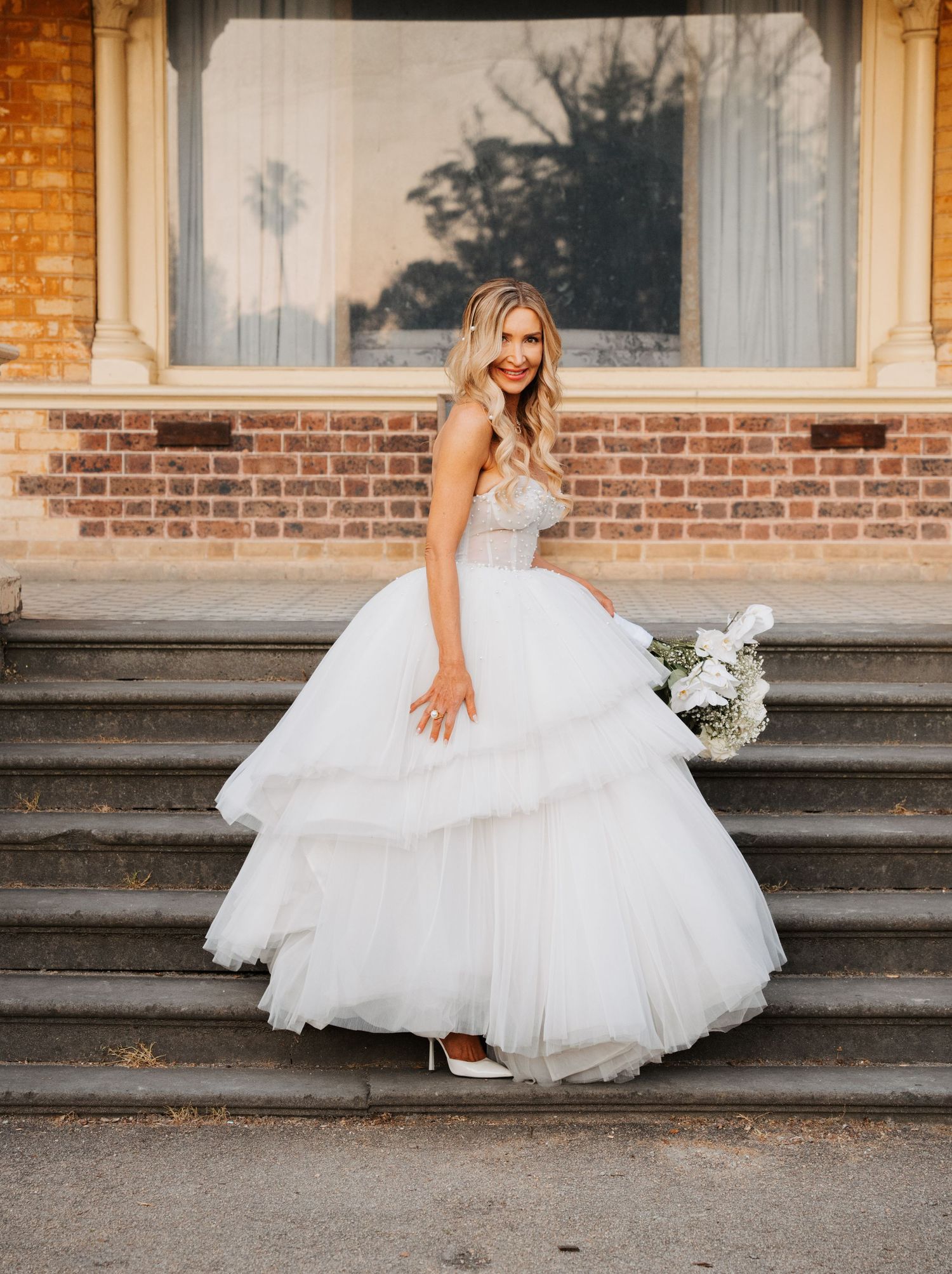 A bride in a layered tulle ball gown poses elegantly on stone steps with a bouquet at sunset.