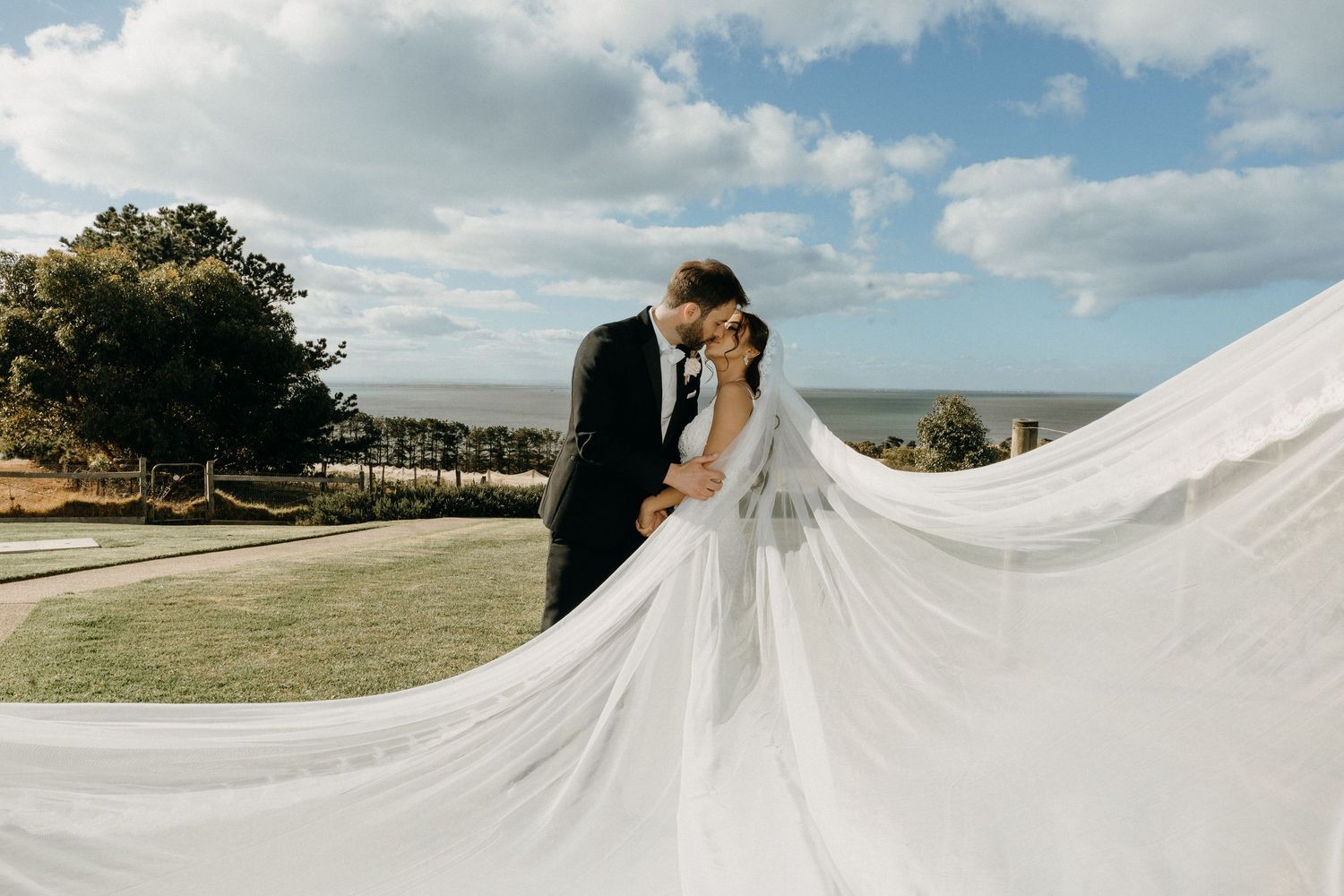 Bride and groom share a romantic kiss outdoors with a flowing white veil billowing in the breeze near the ocean.
