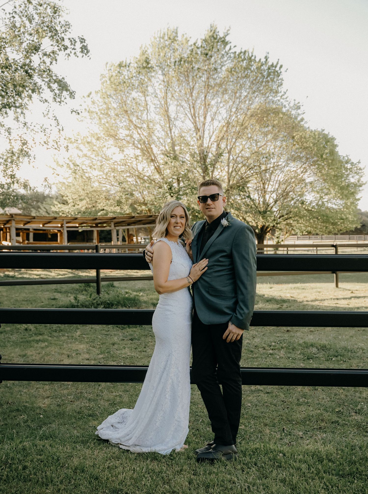 A couple shares romantic moments by a rustic wooden fence during a countryside wedding photoshoot at sunset.