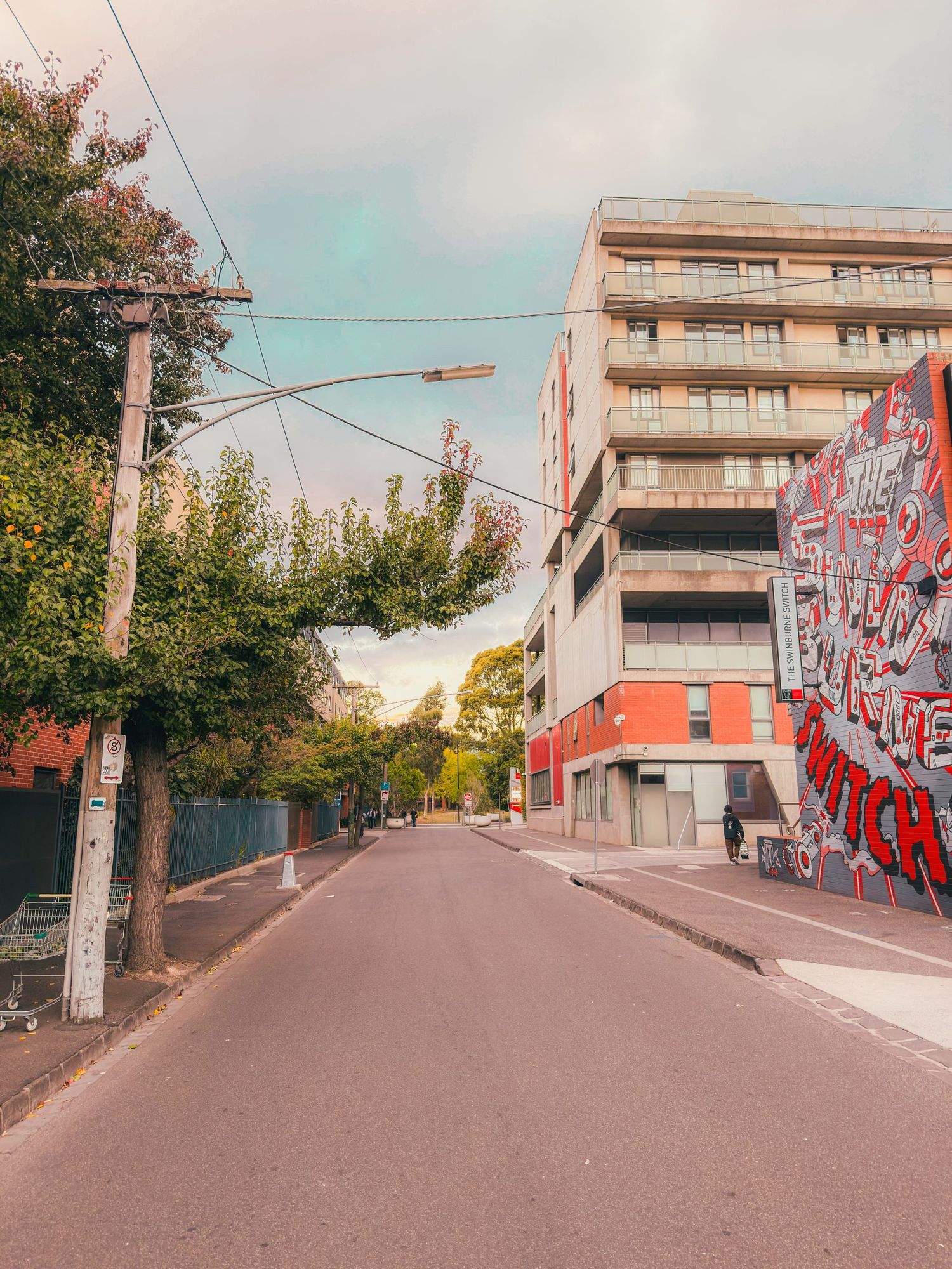 Urban street scene with modern apartment building, colorful graffiti mural, and pink sunset sky in background.