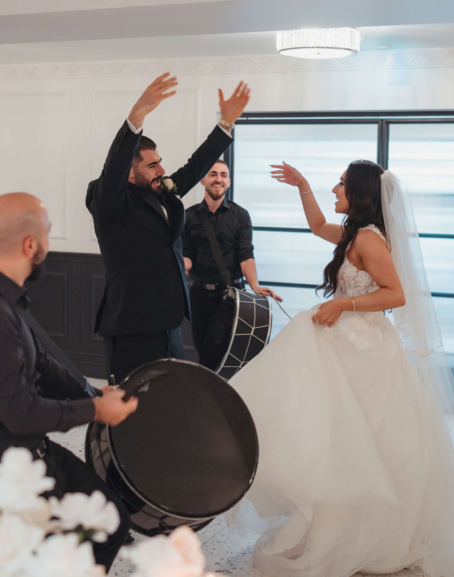 Wedding reception celebration with musicians playing drums while bride in white gown dances excitedly indoors.