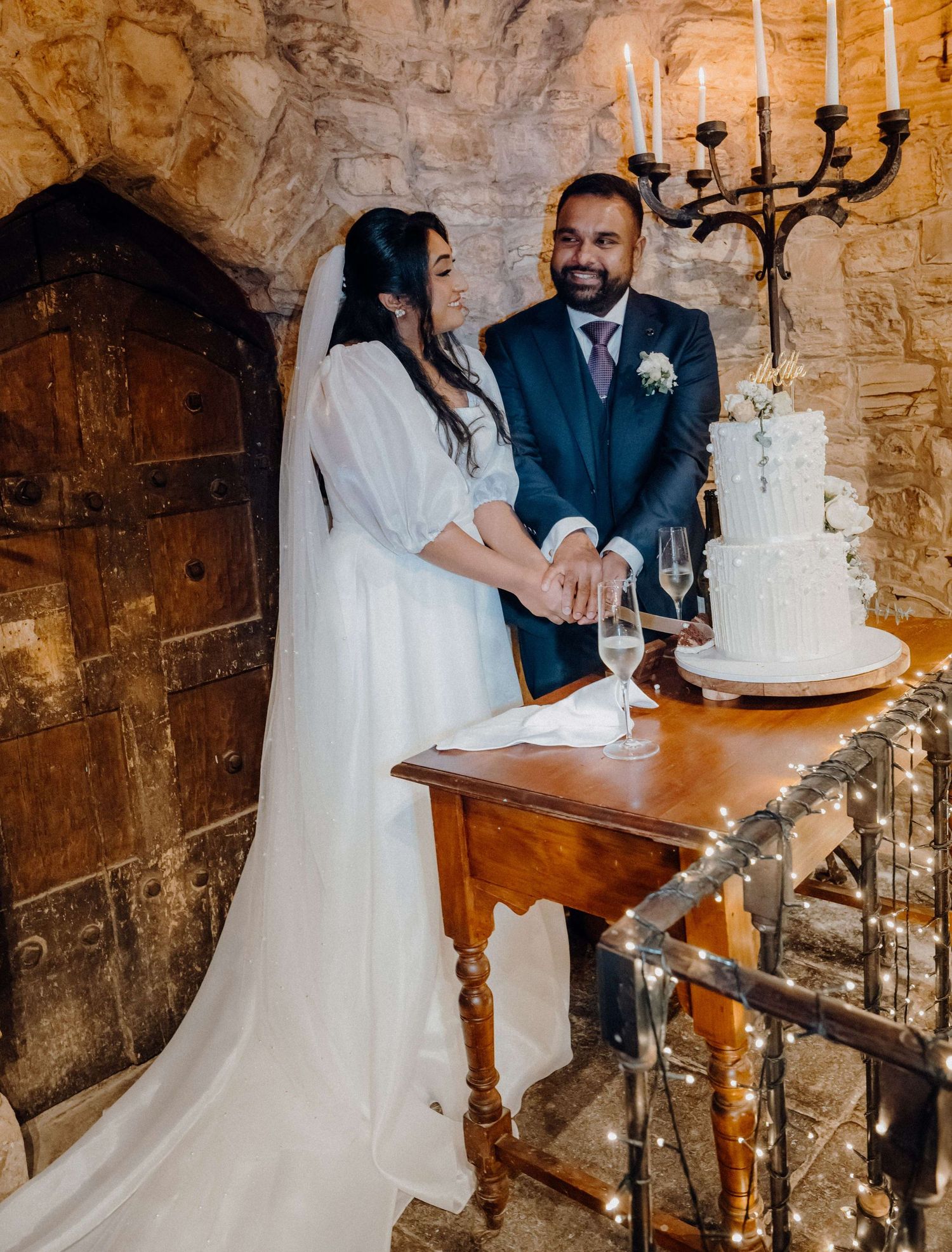 A couple cuts their elegant white wedding cake together in a rustic stone room with a black candelabra.