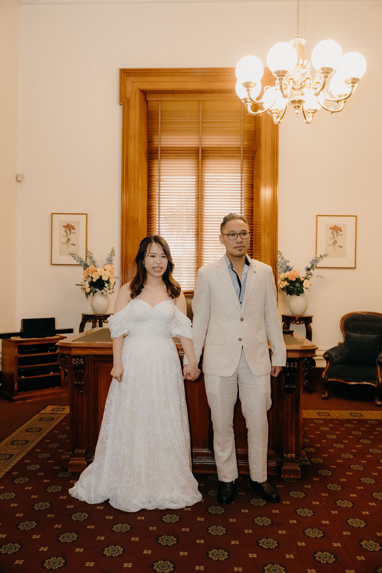 Wedding couple in white dress and light colored suit pose together in an elegant room with patterned carpet and chandeliers.