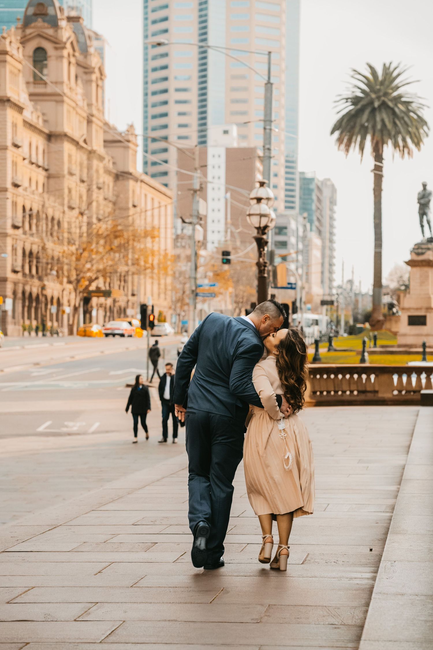 A couple walking together on a wide stone promenade with historic buildings and palm trees in Barcelona, Spain.