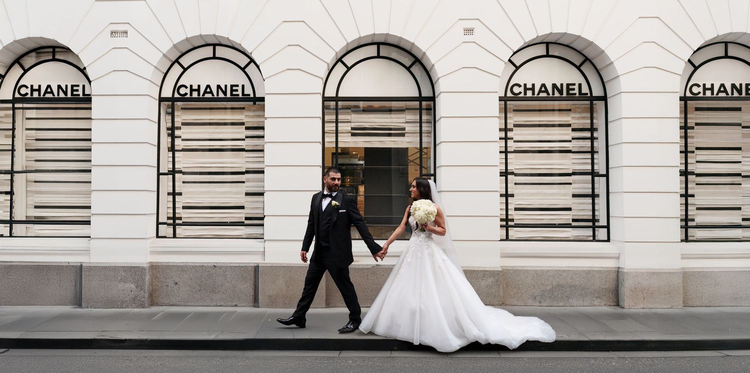 Bride and groom walking hand in hand in front of a Chanel boutique on their wedding day.