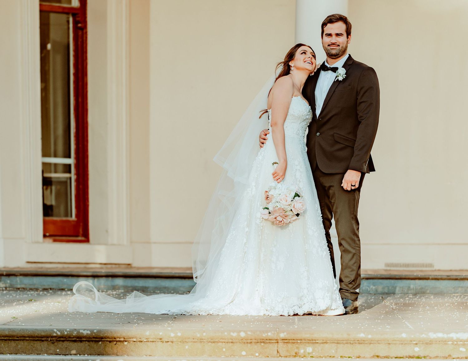 A wedding couple stands together on stone steps wearing elegant attire in soft vintage-style lighting.