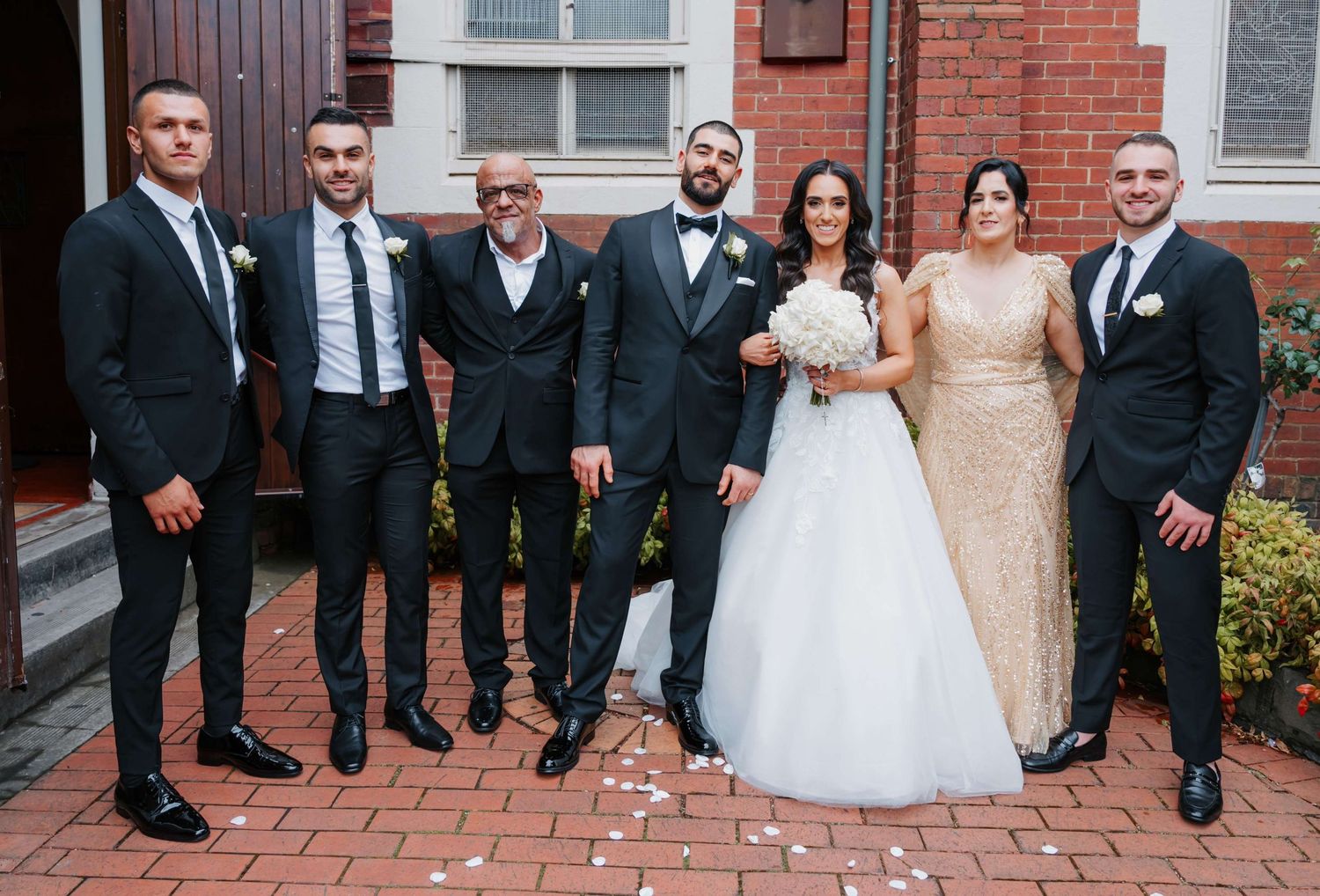 Wedding party in formal attire poses together on brick pathway outside historic building.