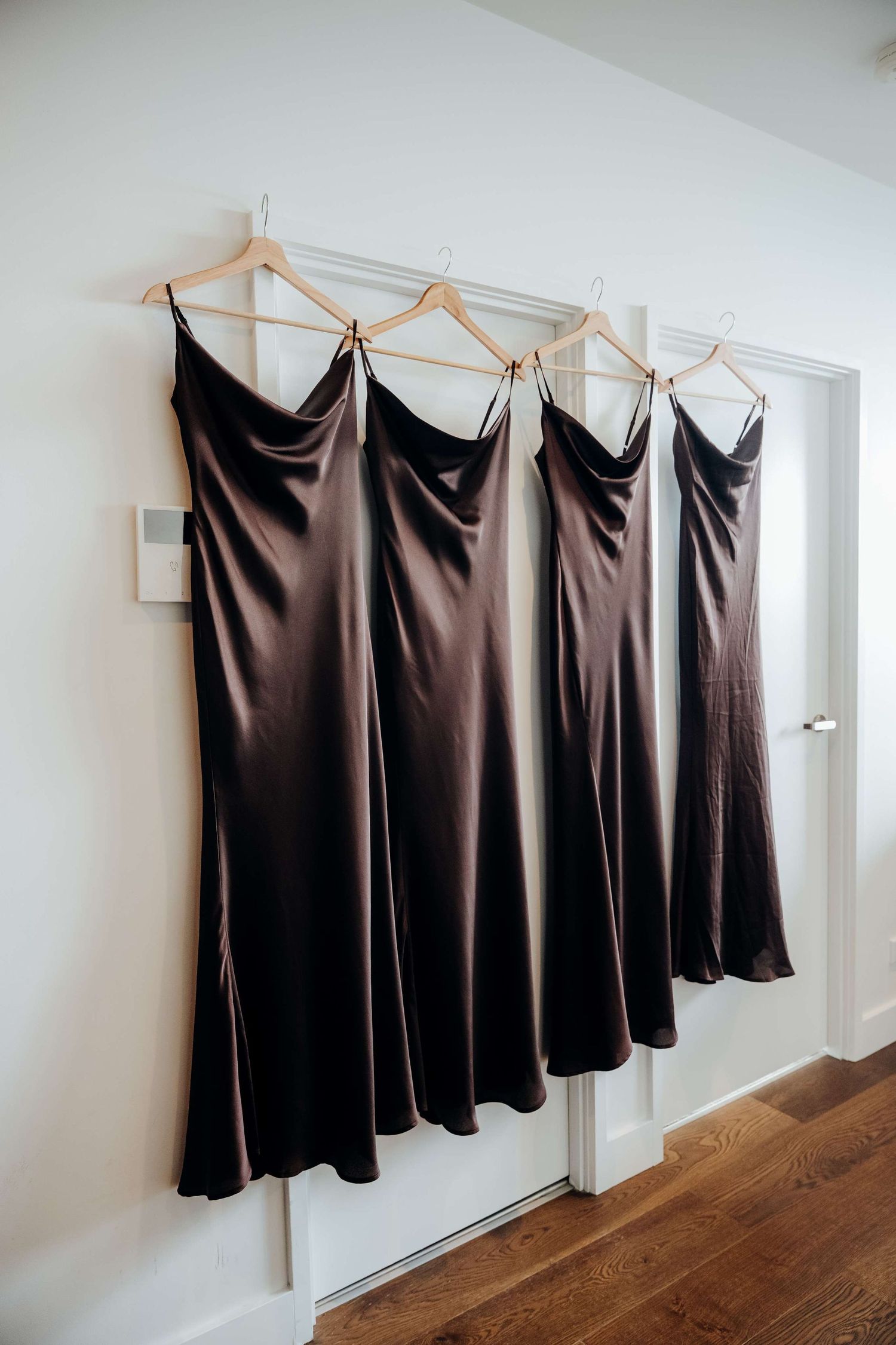 Different views of a row of black satin dresses hanging on wooden hangers against a white wall with hardwood floors.