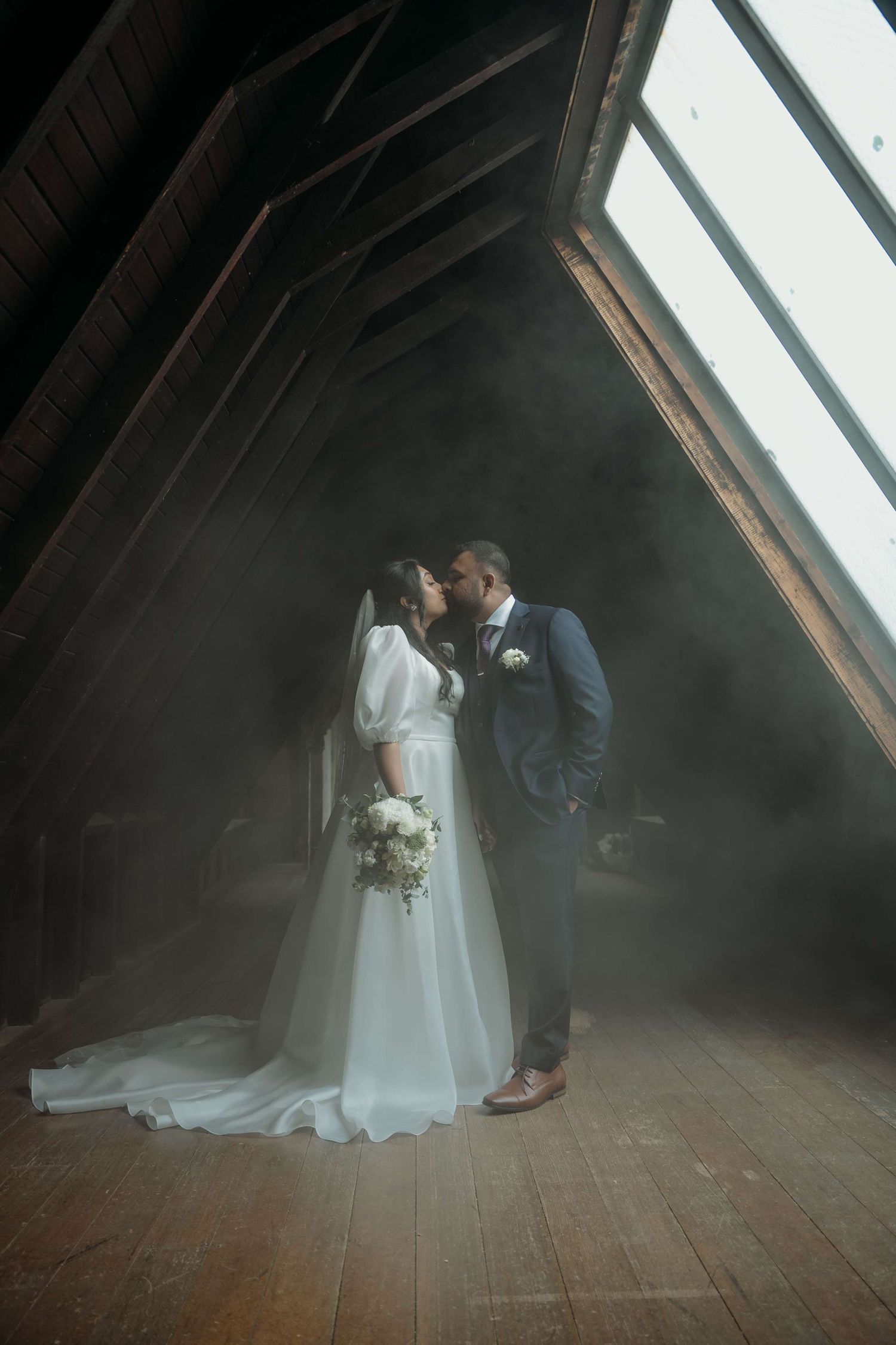 A romantic wedding scene shows a couple embracing beside angled windows in a rustic attic setting with natural lighting.