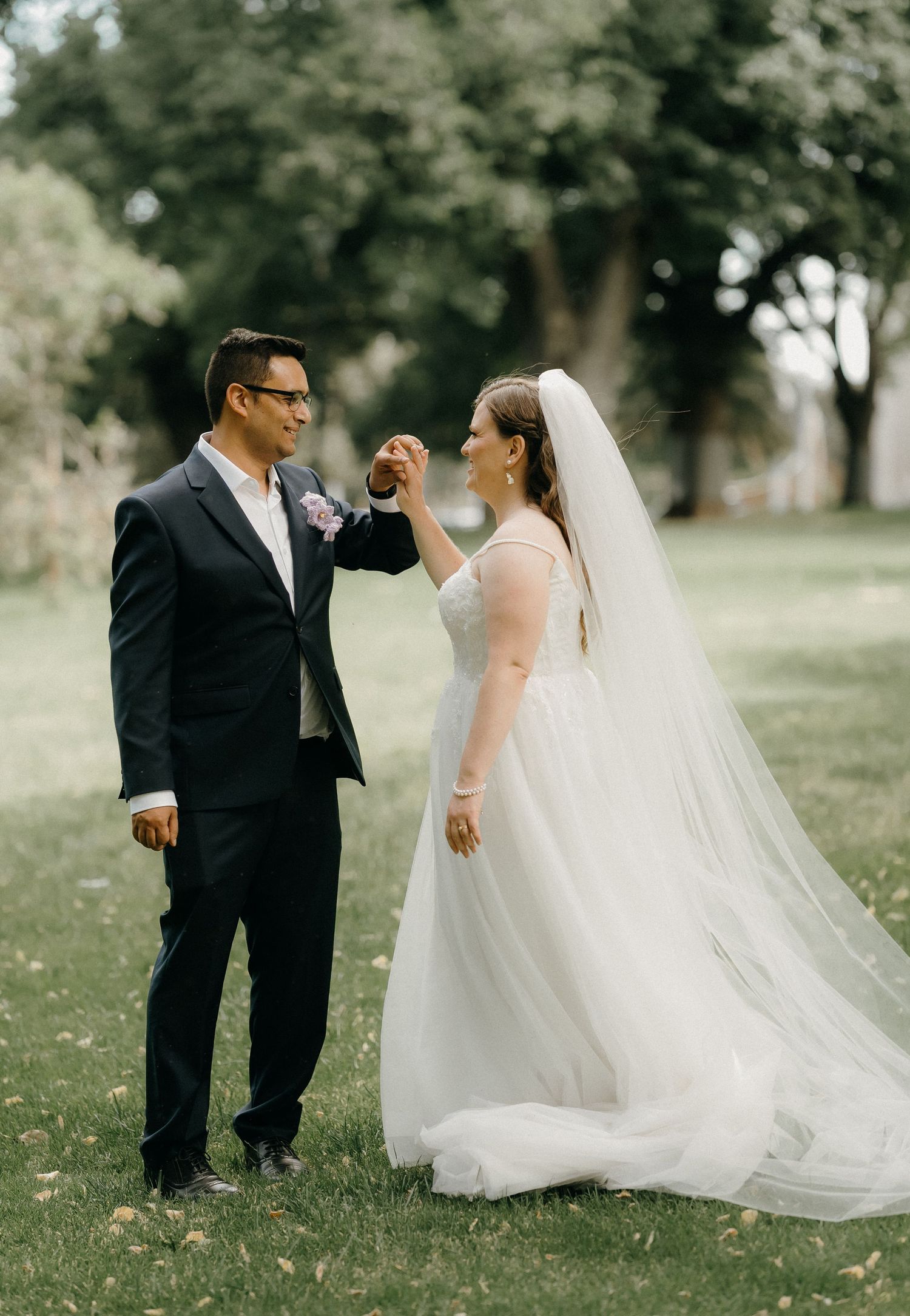 A romantic outdoor wedding photo showing a couple sharing a joyful moment on a grassy lawn with trees in the background.