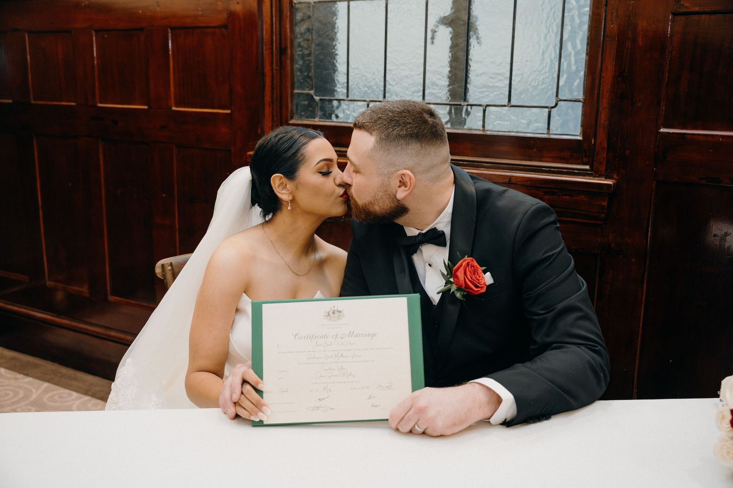 Bride and groom kissing while holding marriage certificate at wedding signing table.