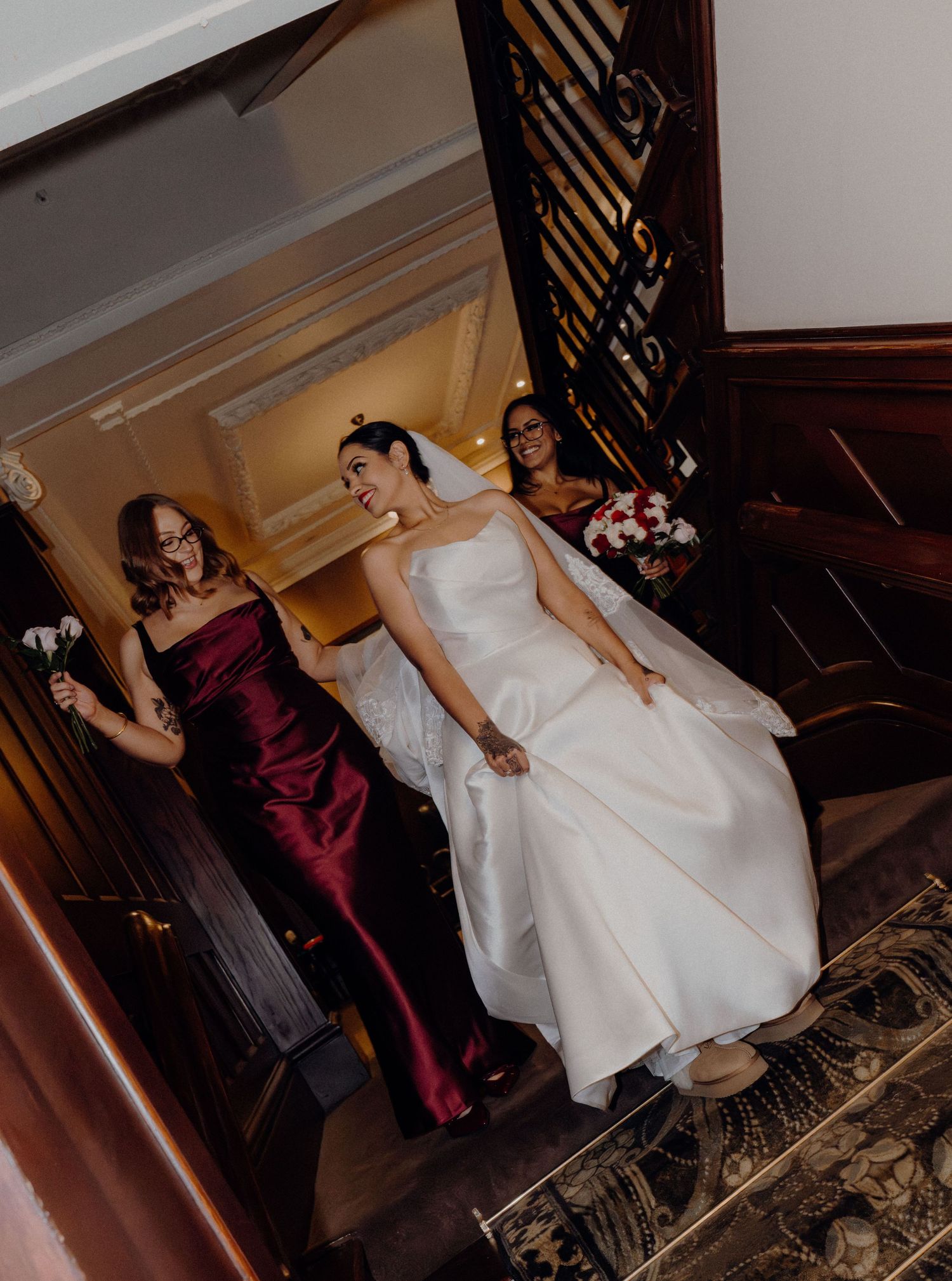 Bride in white gown descends ornate staircase with bridesmaids in burgundy dresses holding bouquets at wedding venue.