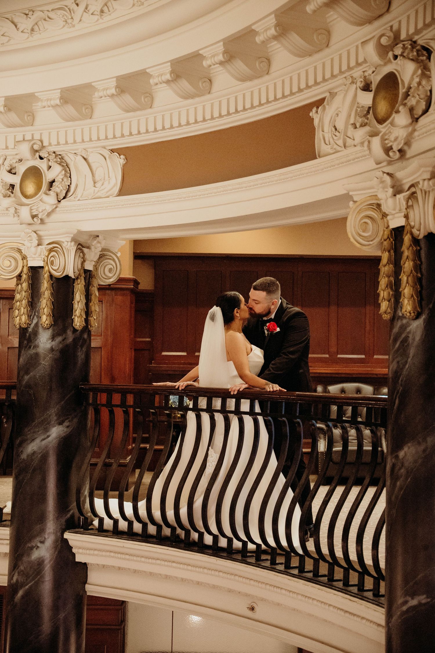 Bride and groom share a romantic kiss on an ornate balcony with marble columns and gold accents in an elegant venue.