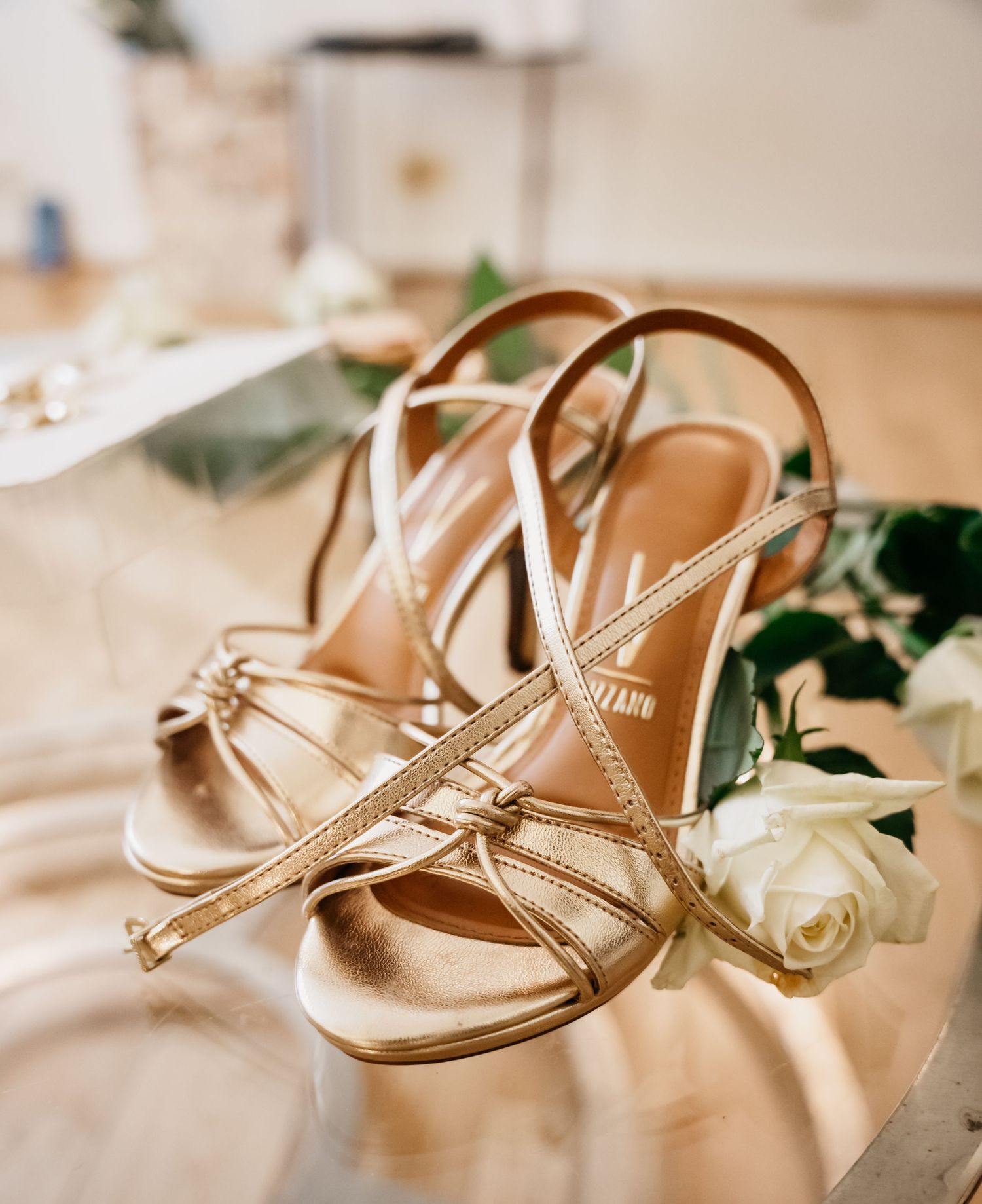 Elegant metallic gold bridal sandals with strappy design displayed next to a white rose on a sunlit surface.