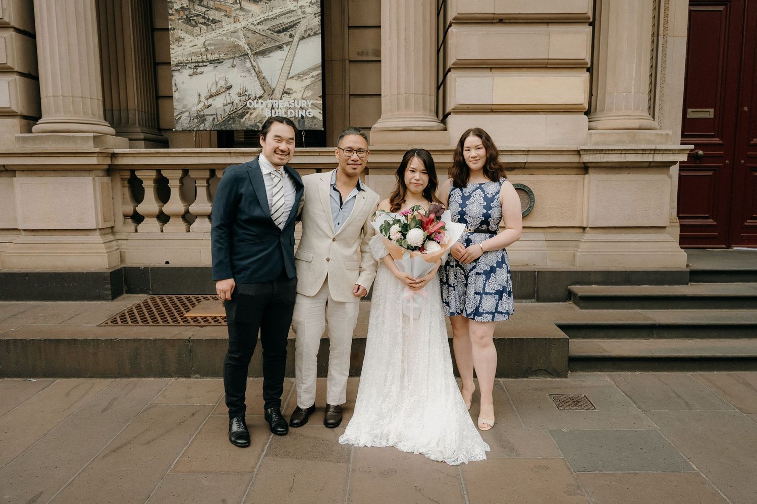 A wedding party poses together on grand stone steps of a historic building in elegant formal attire.