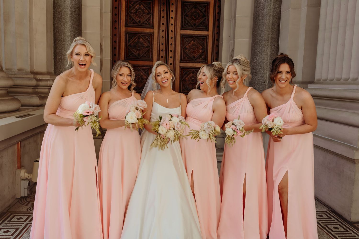 Bridesmaids in matching pink dresses pose with bride in white gown at elegant venue with classical columns.