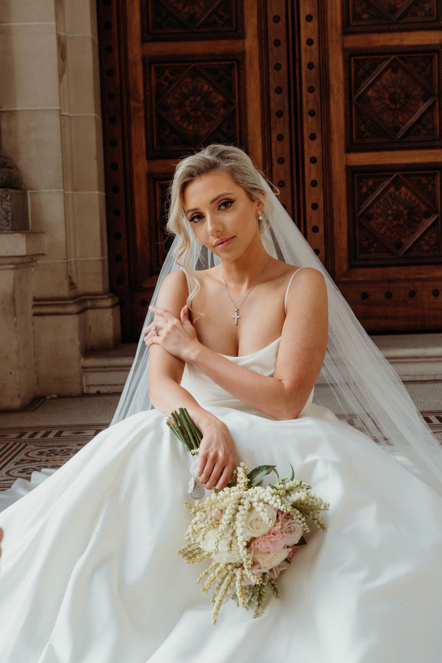 A bride in a white wedding gown poses elegantly against ornate wooden doors while holding a romantic bouquet.