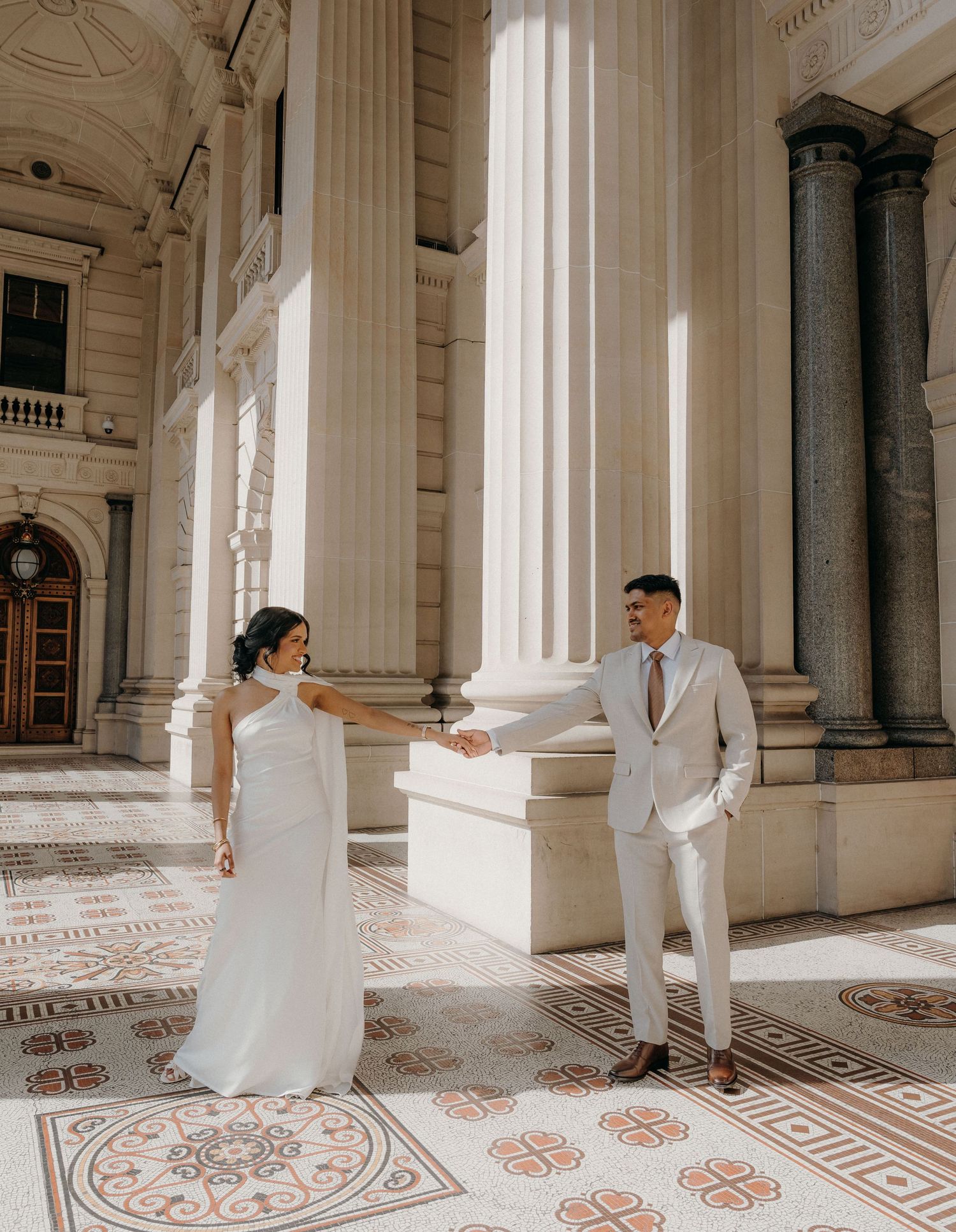 Elegant couple in formal attire holding hands on ornate marble floor with classical columns in the background.