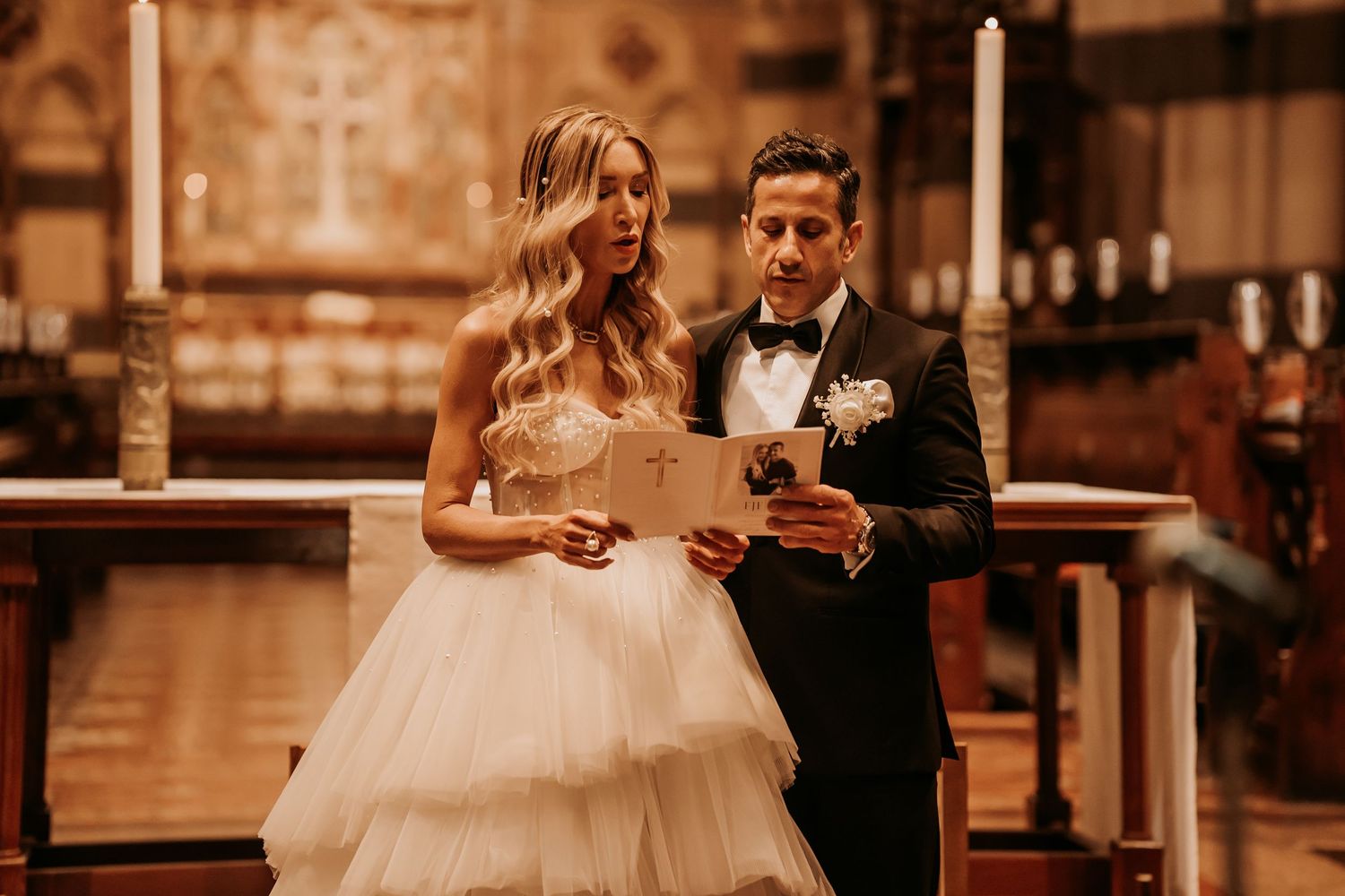 A bride in a layered tulle gown and groom in tuxedo share a moment reading documents during their church ceremony.