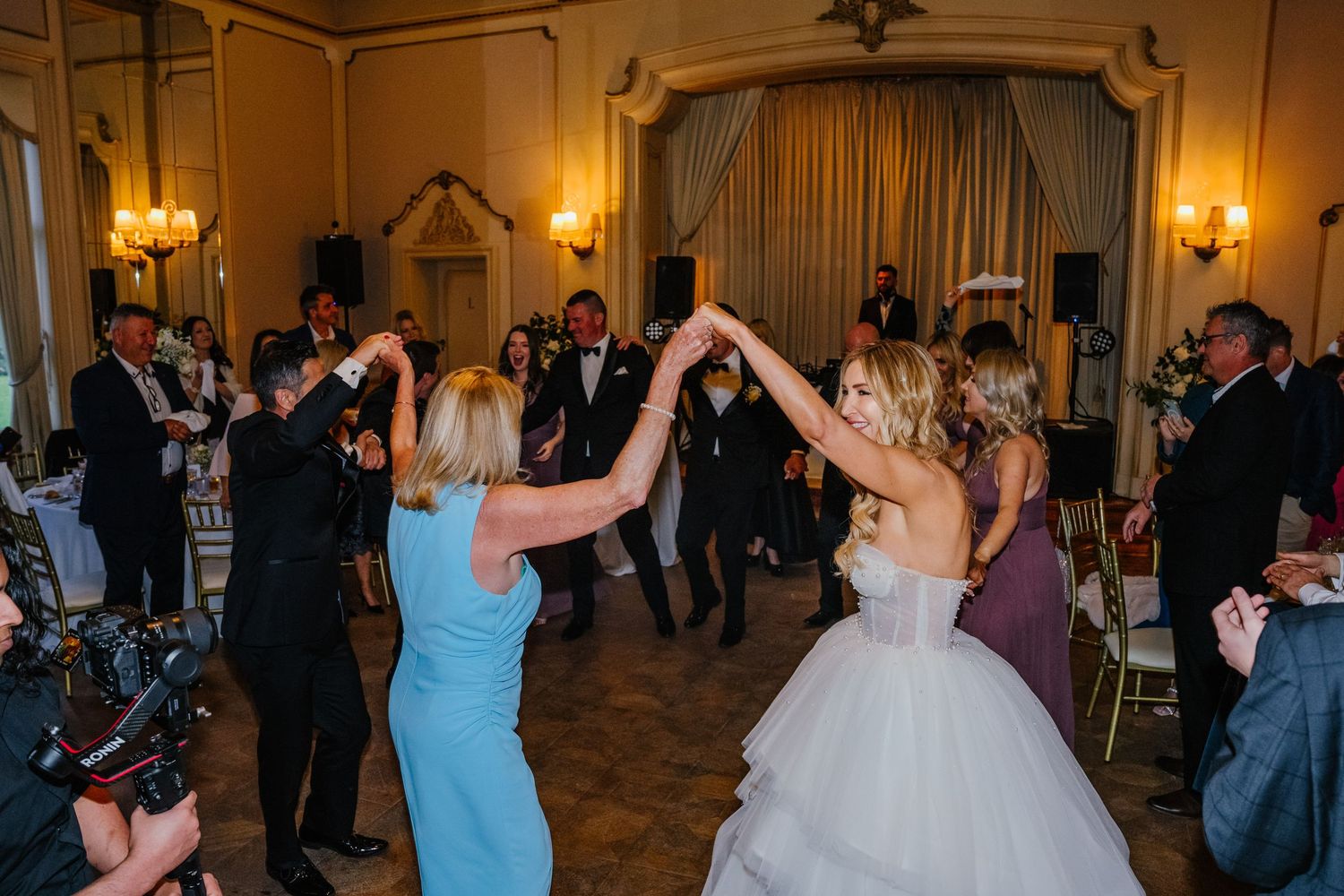 Wedding guests dance and celebrate together in an elegant ballroom with warm lighting and ornate decor.