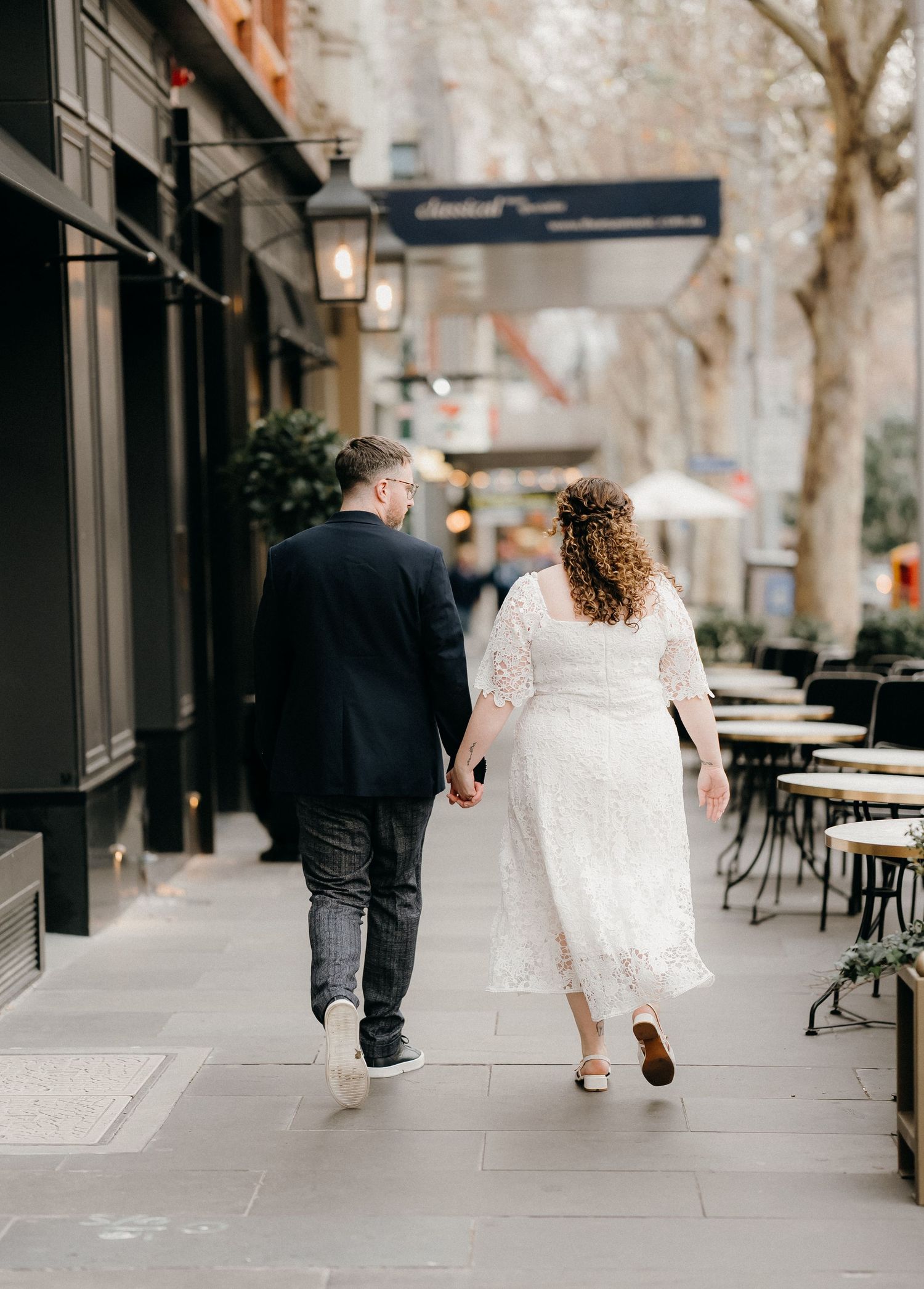 A couple walking together on a city sidewalk with outdoor cafe seating visible in a romantic urban setting.